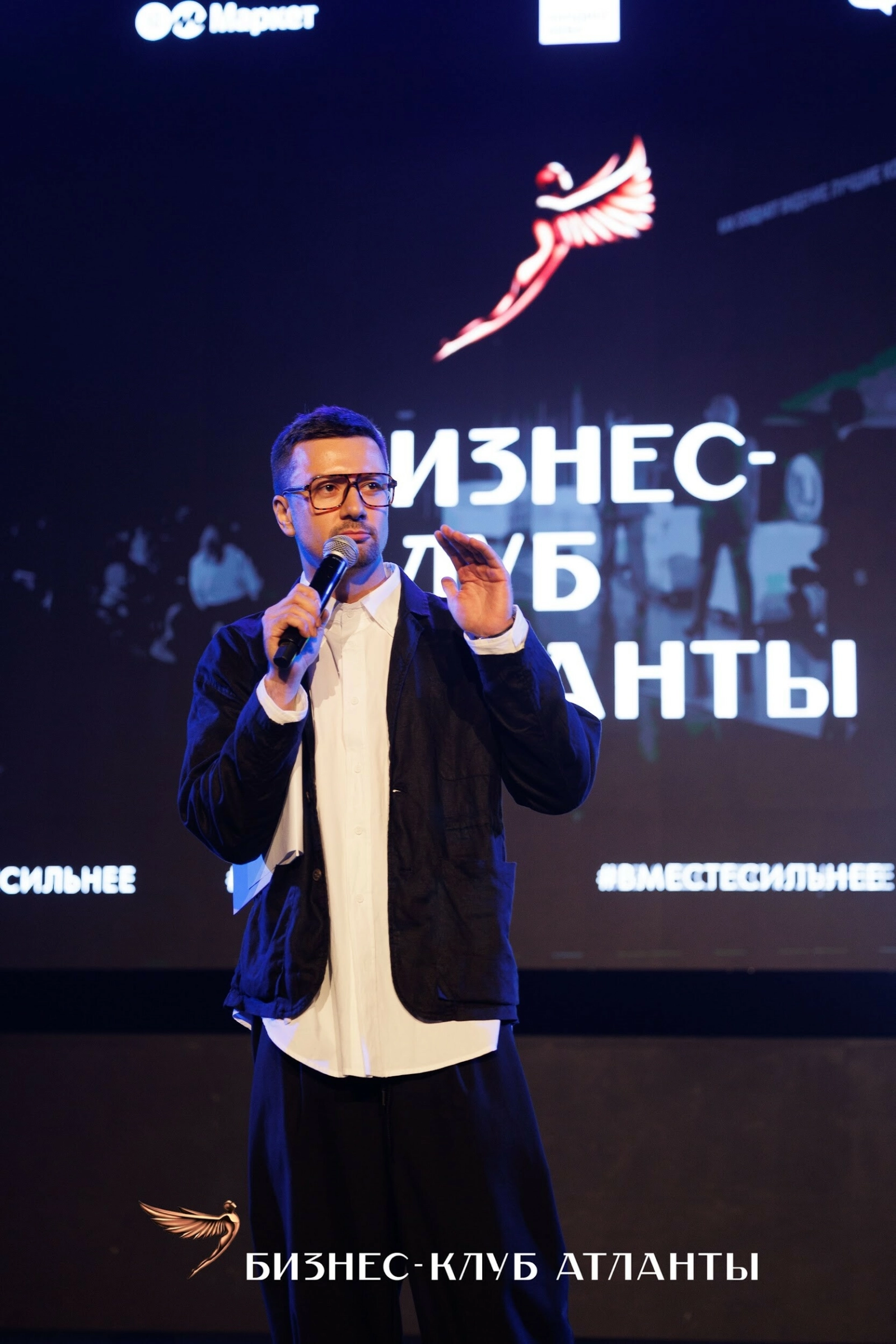 Ведущий в очках жестикулирует с микрофоном на фоне экрана форума