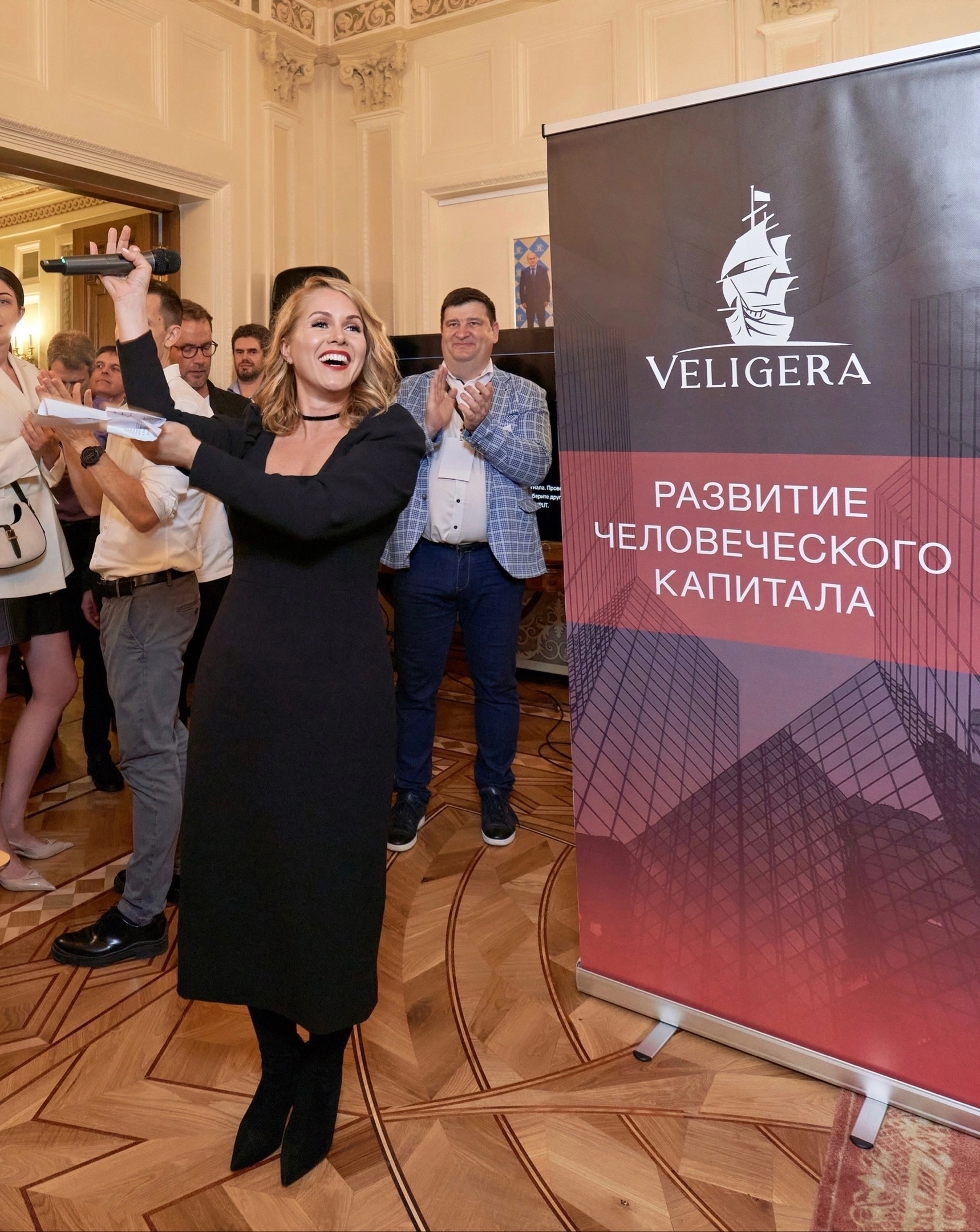 ведущая поднимает микрофон на фоне баннера мероприятия перед гостями