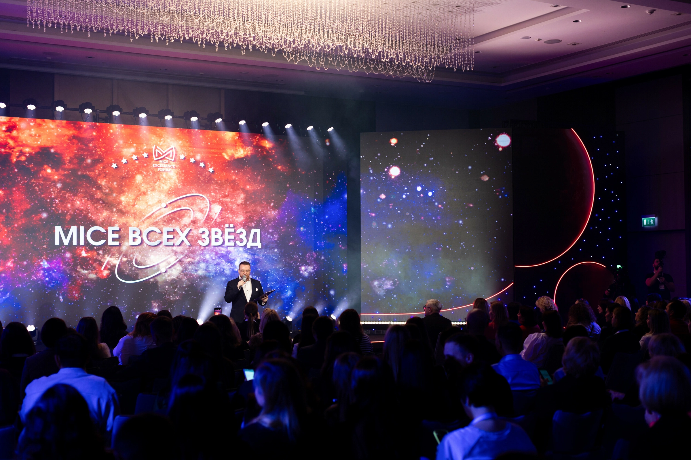 ведущий на сцене на фоне звёздного экрана перед залом гостей