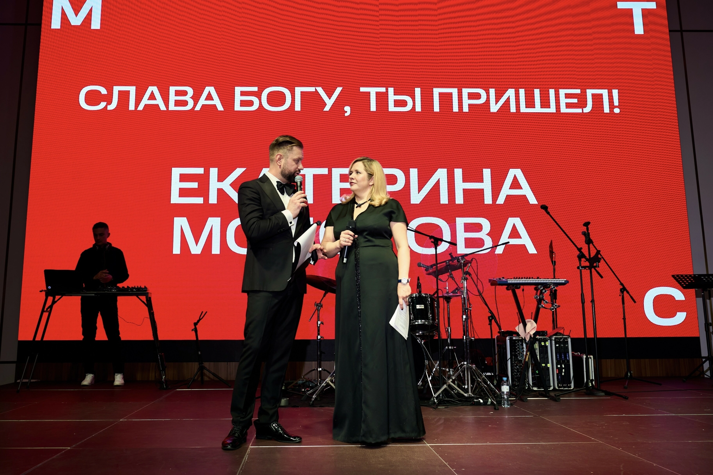 ведущий и женщина с микрофонами стоят на сцене у красного экрана