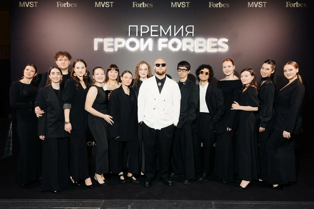 группа людей в чёрных нарядах на фотозоне премии герой forbes с логотипами на фоне