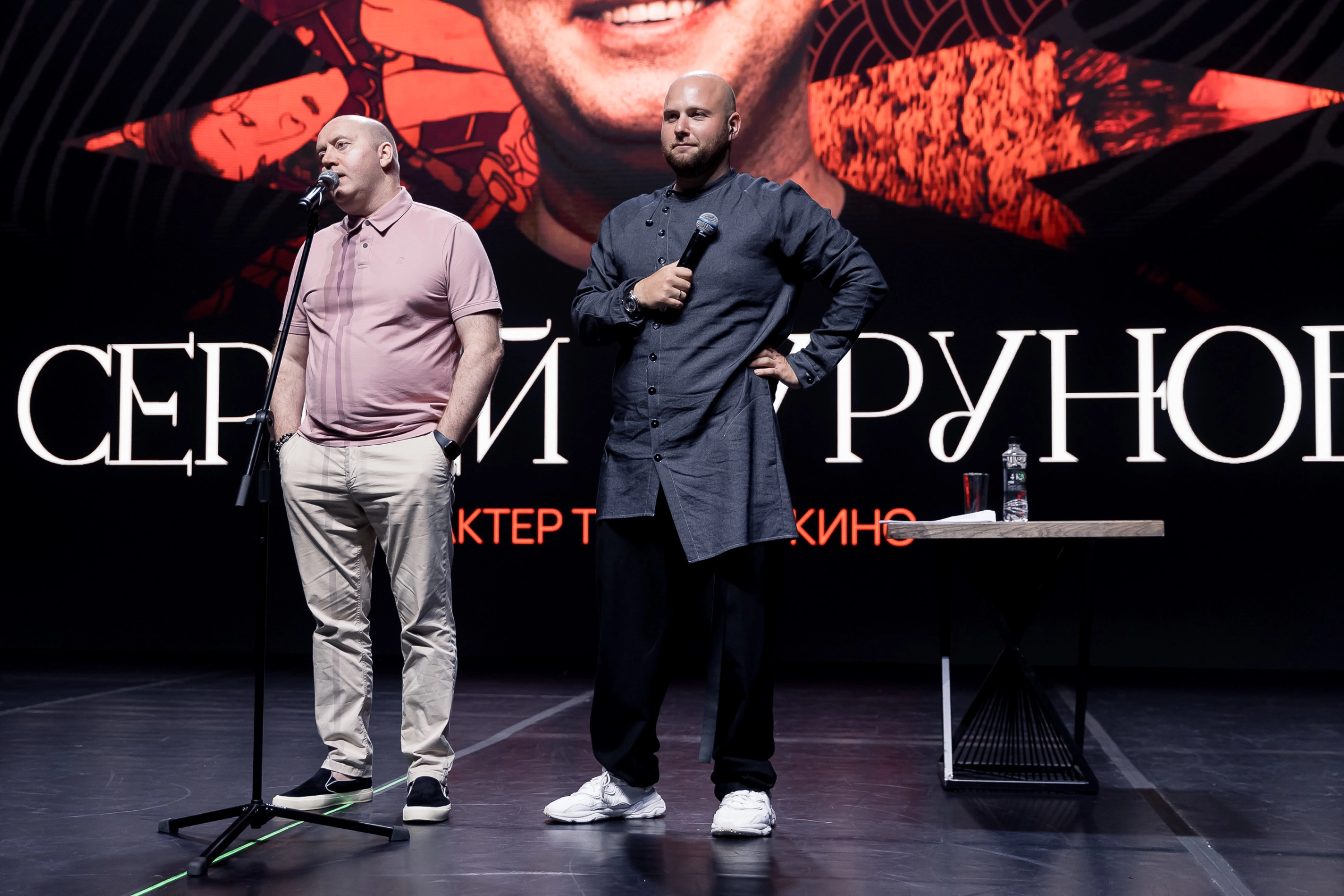 Два ведущих с микрофонами на сцене перед экраном во время творческого вечера