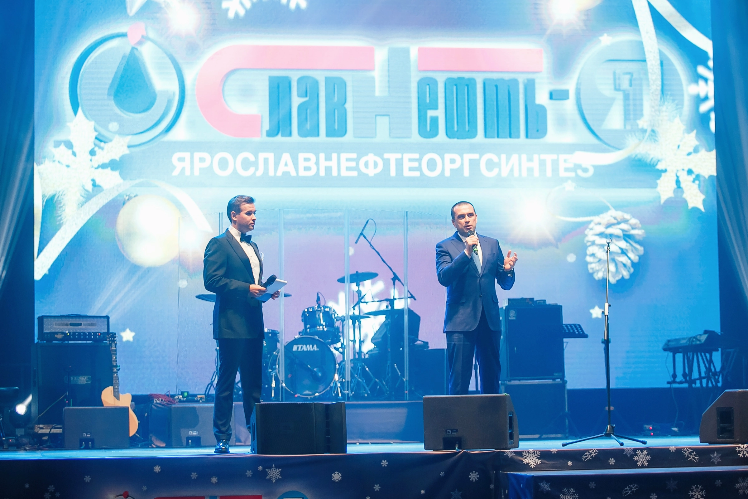 Двое ведущих в костюмах на сцене с новогодним фоном компании