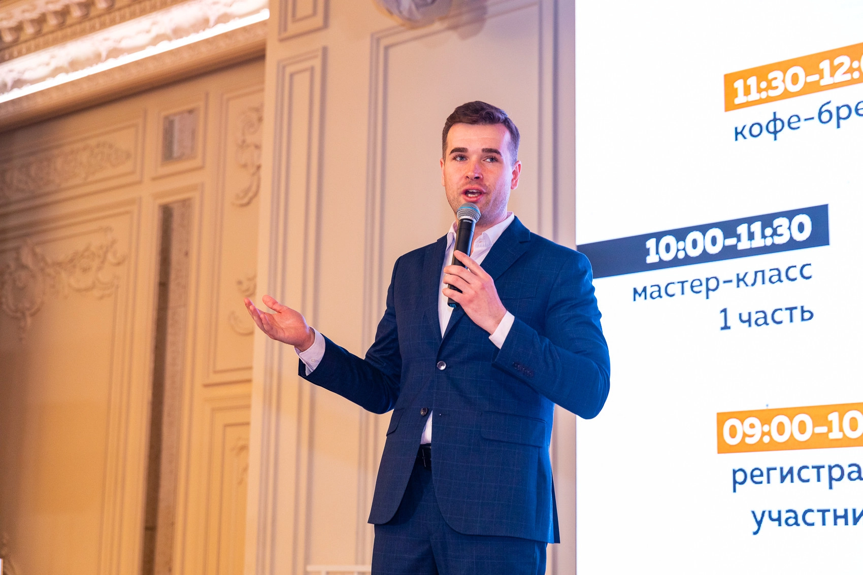 Мужчина в костюме с микрофоном на фоне экрана с расписанием