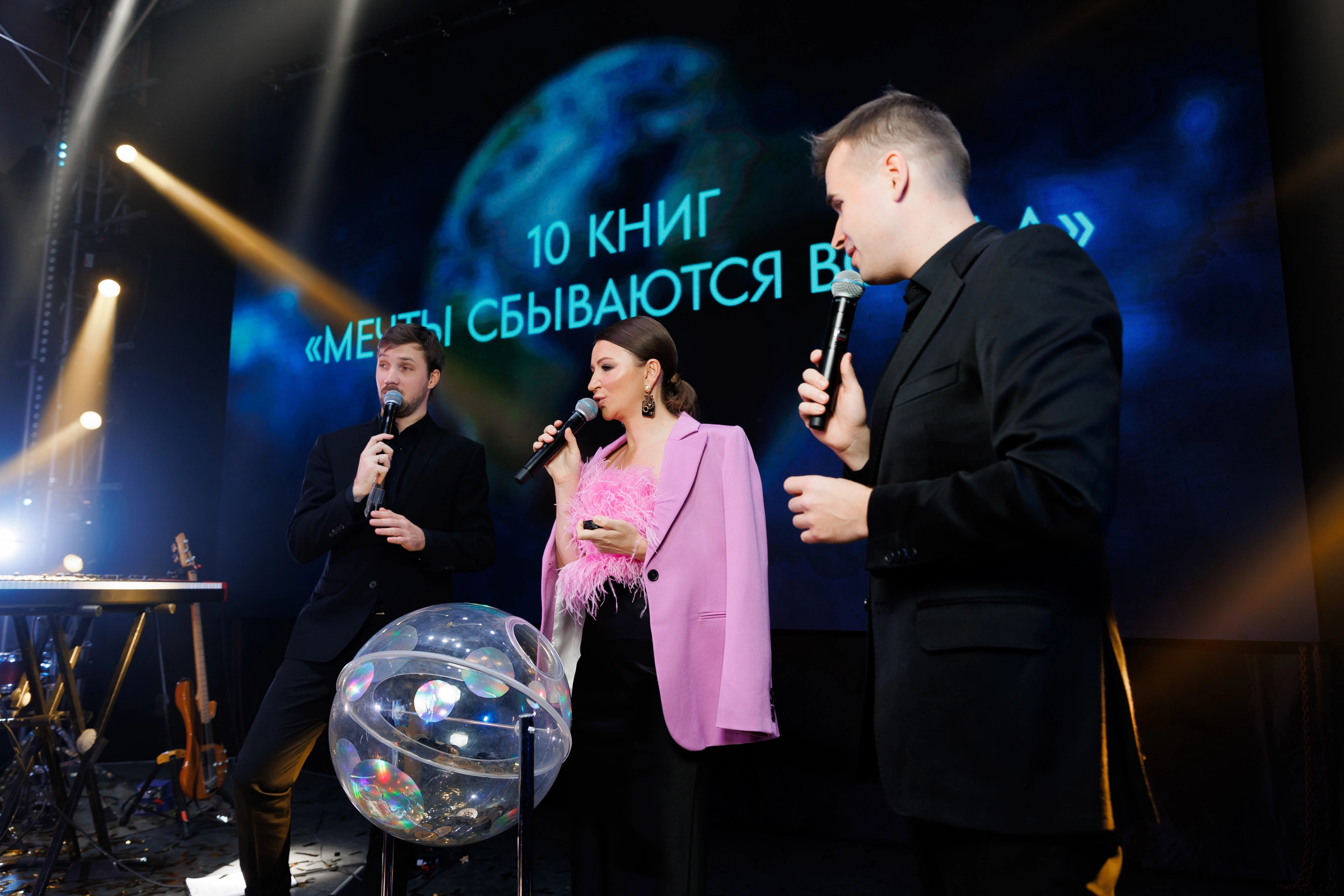 Трое ведущих с микрофонами на сцене на фоне яркого экрана