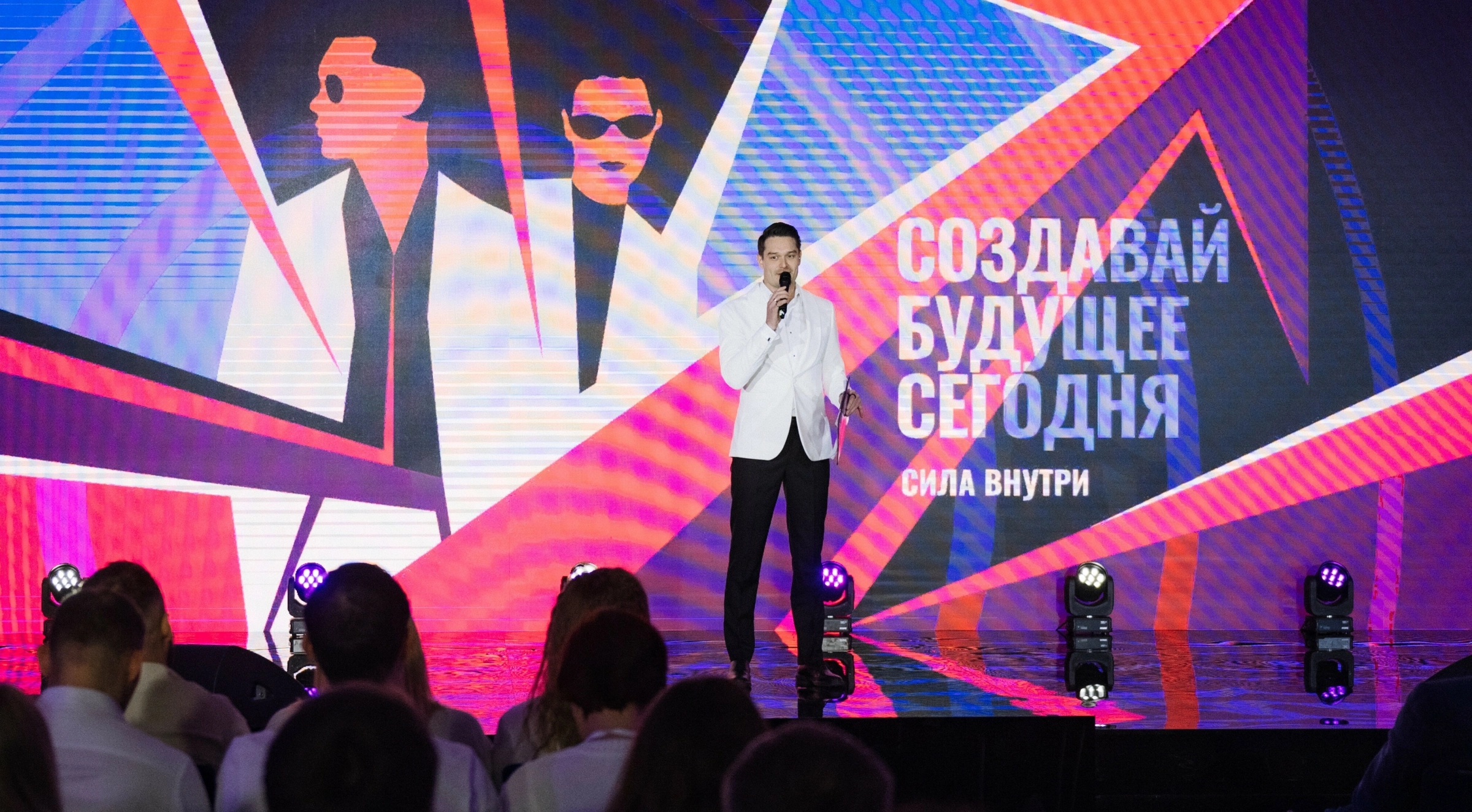 ведущий в белом костюме на сцене с яркой неоновой графикой