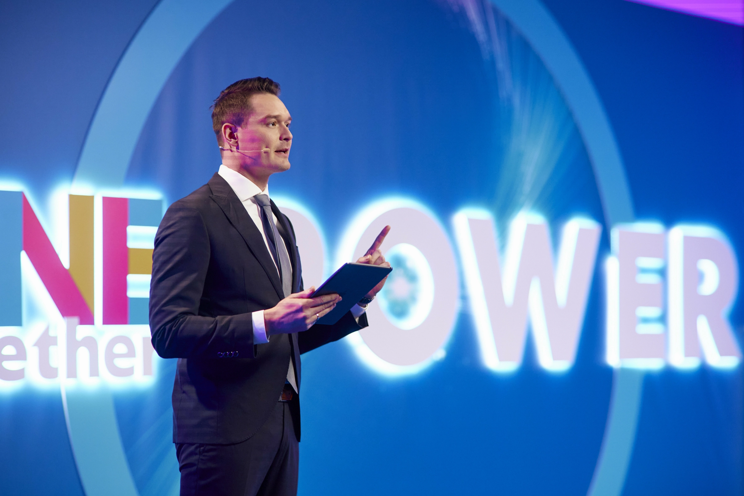 Ведущий с планшетом на сцене на фоне светящейся надписи Power