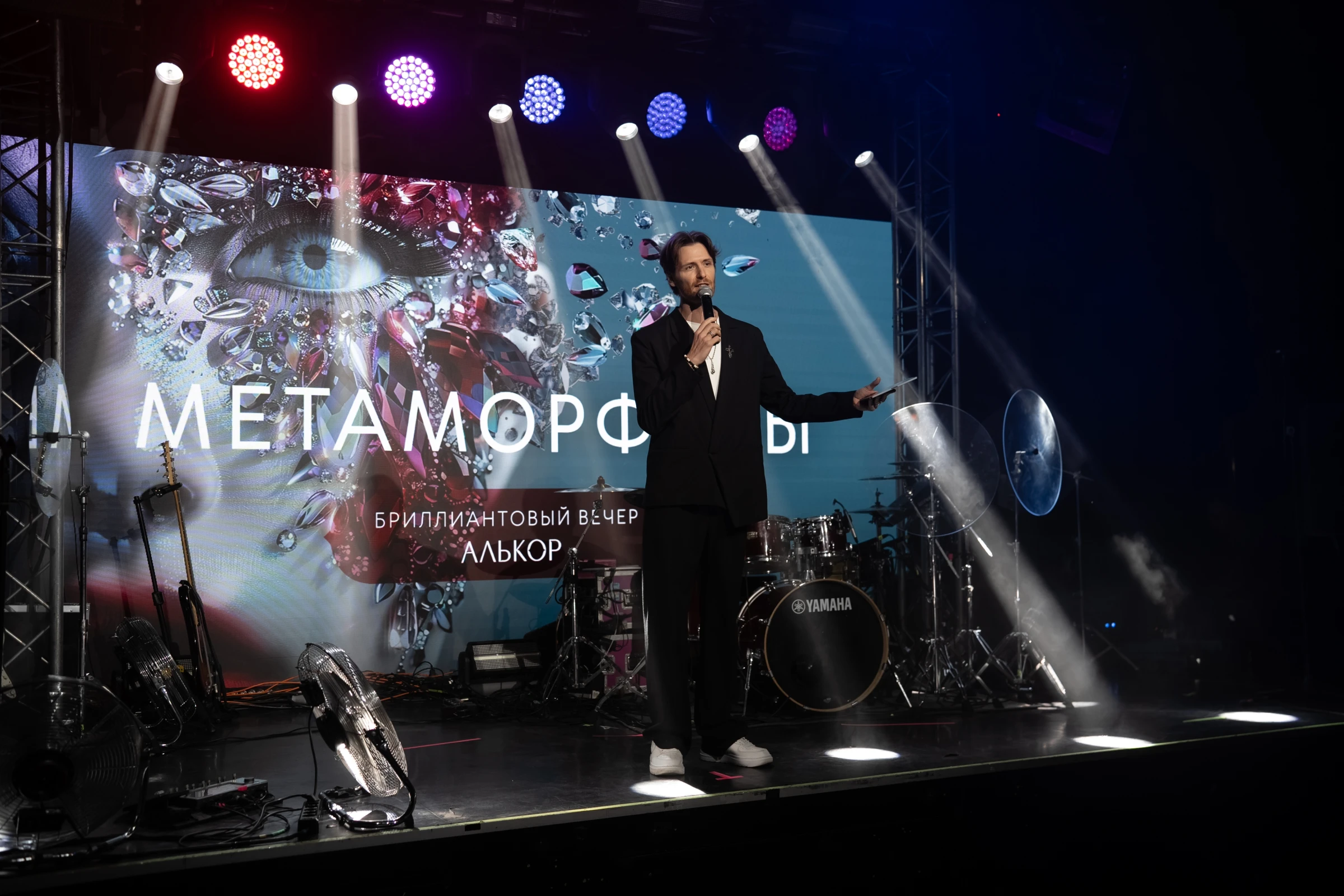 Ведущий с микрофоном на сцене под лучами прожекторов у большого экрана