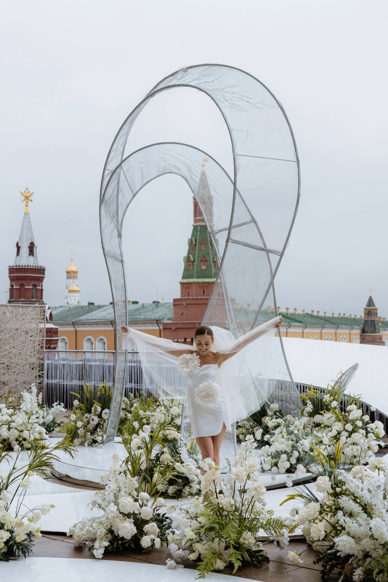 Невеста в коротком платье среди белых цветов и скульптурных арок на крыше