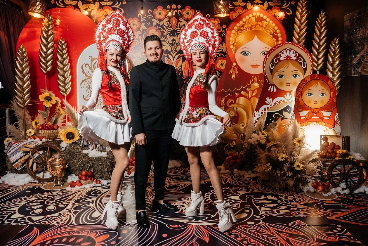 Ведущий с артистками в кокошниках на фоне декора с гигантскими матрёшками