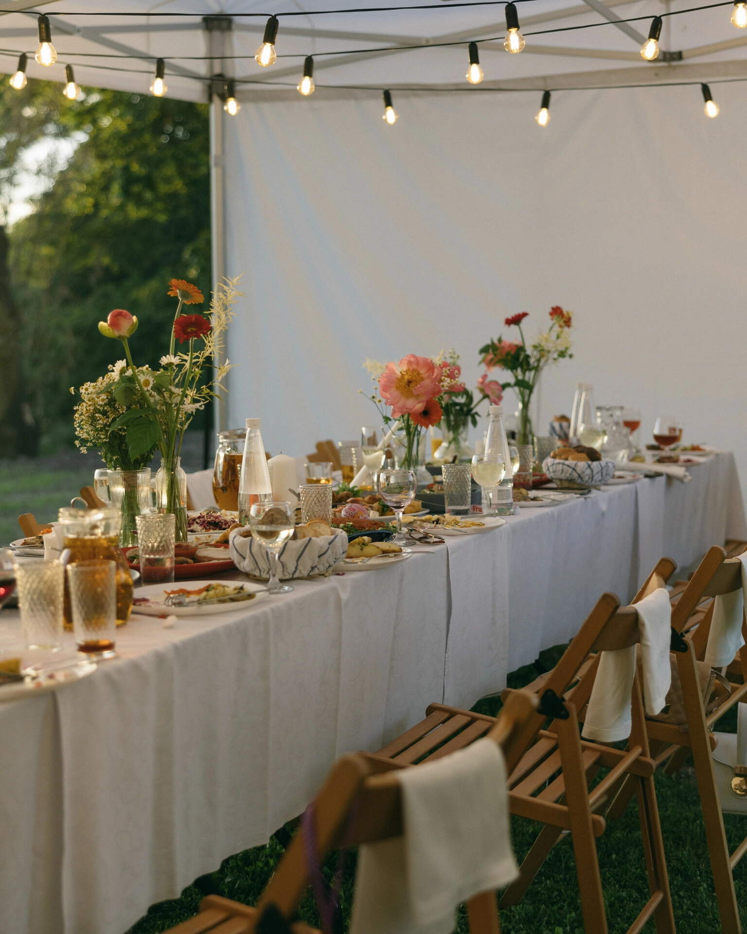 Длинный стол свадебного банкета под шатром с гирляндой лампочек