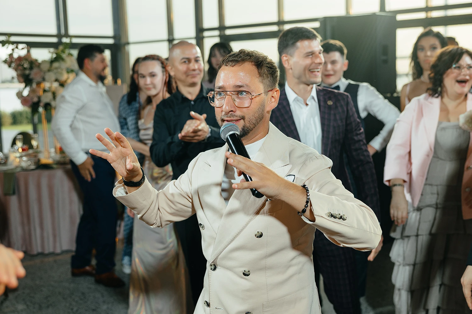 Ведущий свадьбы с микрофоном в бежевом костюме перед смеющимися гостями в банкетном зале