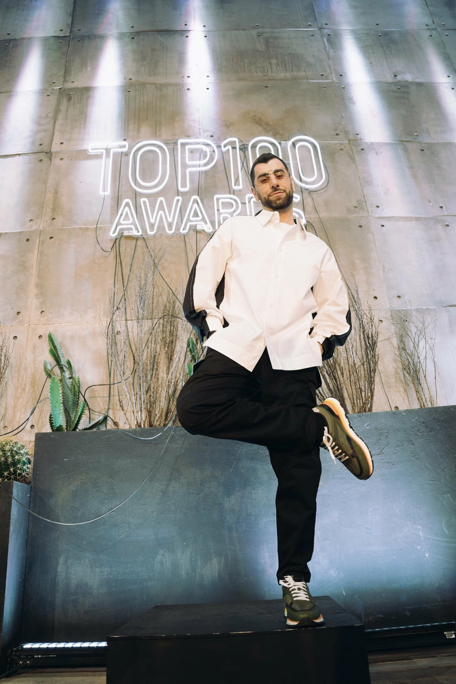 ведущий в белой рубашке позирует у неоновой вывески TOP 100 Awards на премии