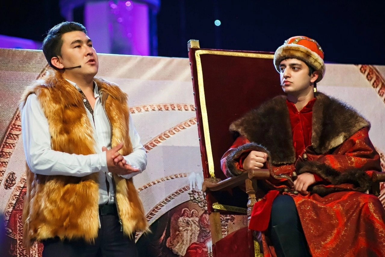ведущий в меховом жилете с микрофоном выступает на сцене рядом с партнёром в костюме царя