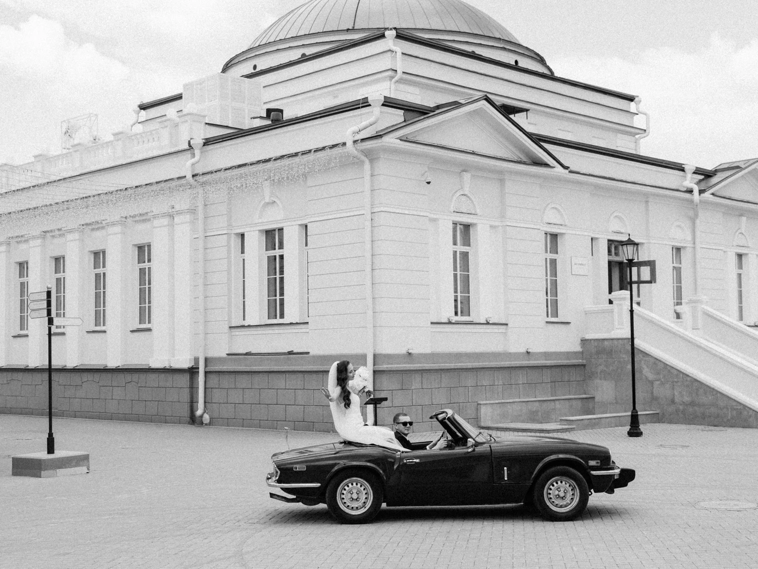 молодожёны едут в чёрном ретро-кабриолете возле старинного исторического здания