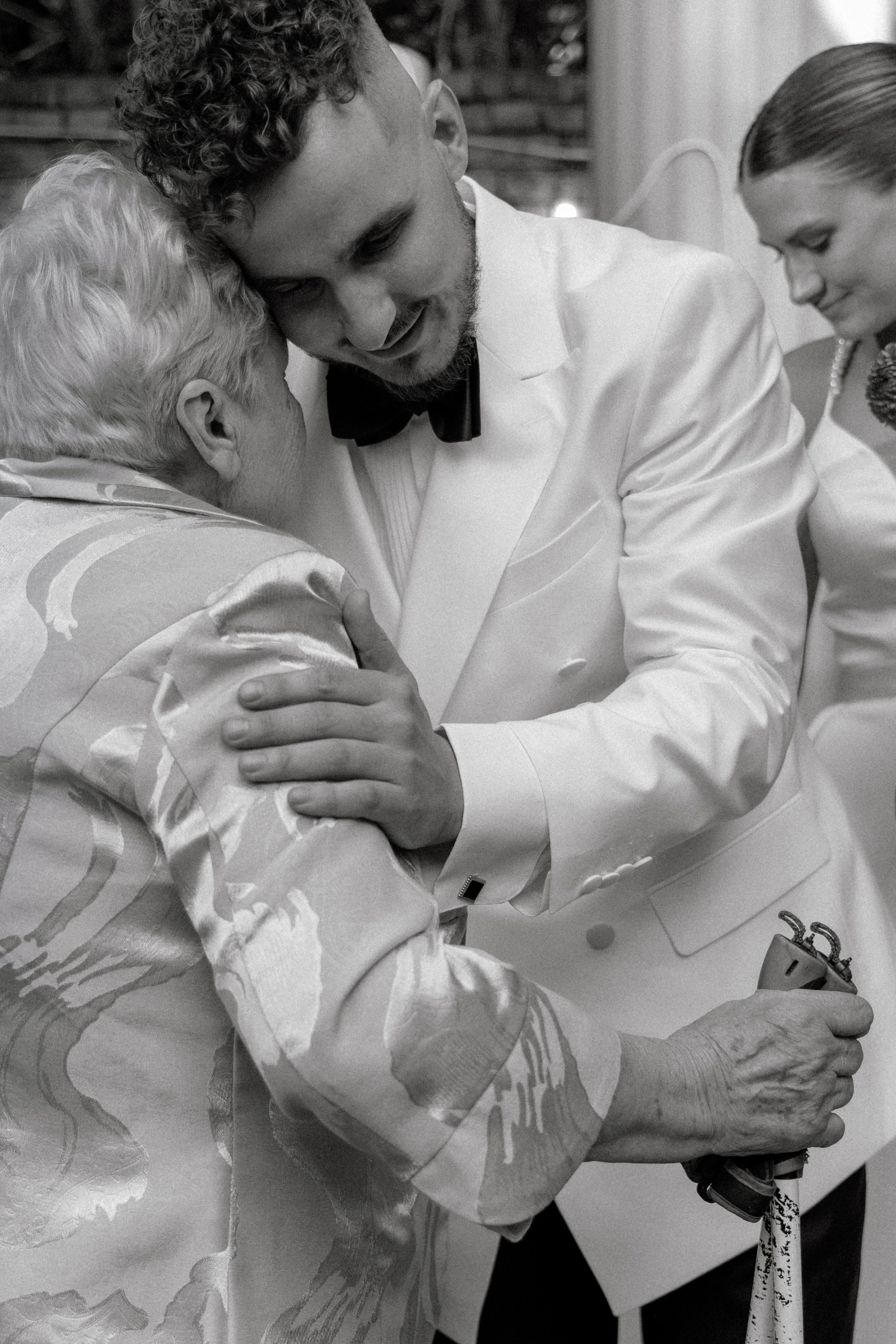 жених в белом смокинге нежно обнимает пожилую родственницу невесты