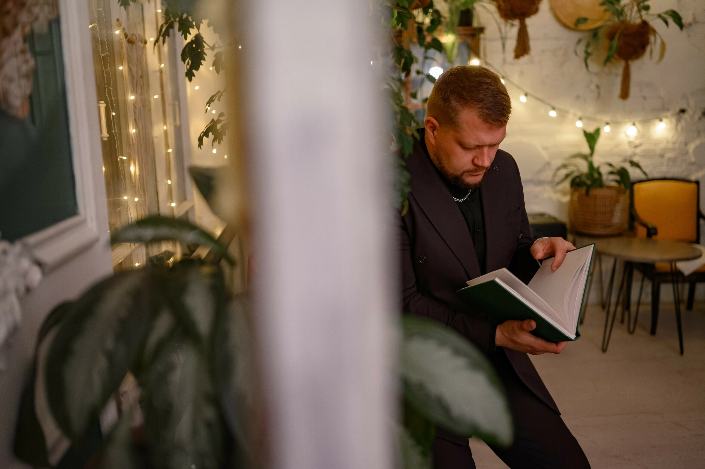 ведущий с книгой в руках в тёплом интерьере с гирляндами и растениями