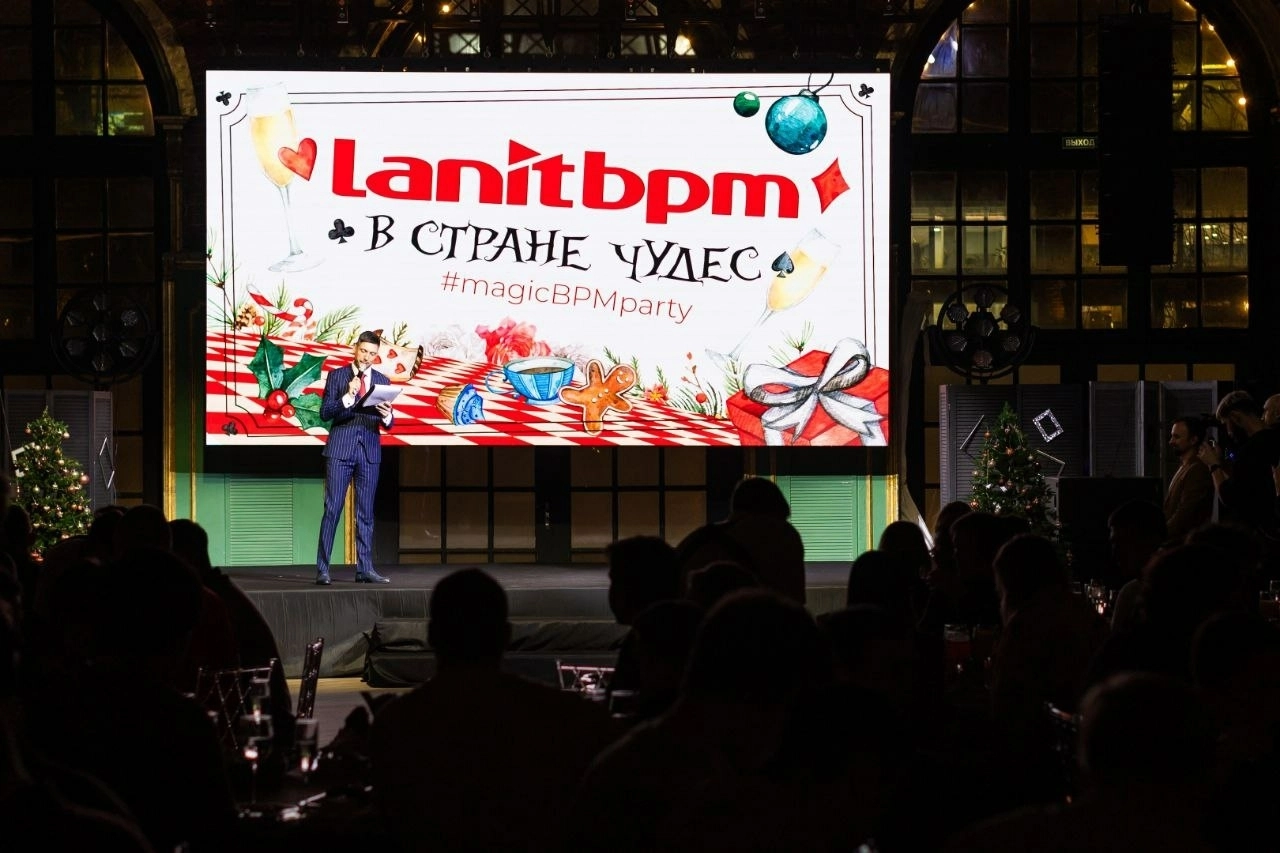 Ведущий с микрофоном стоит на сцене на фоне праздничного экрана вечера