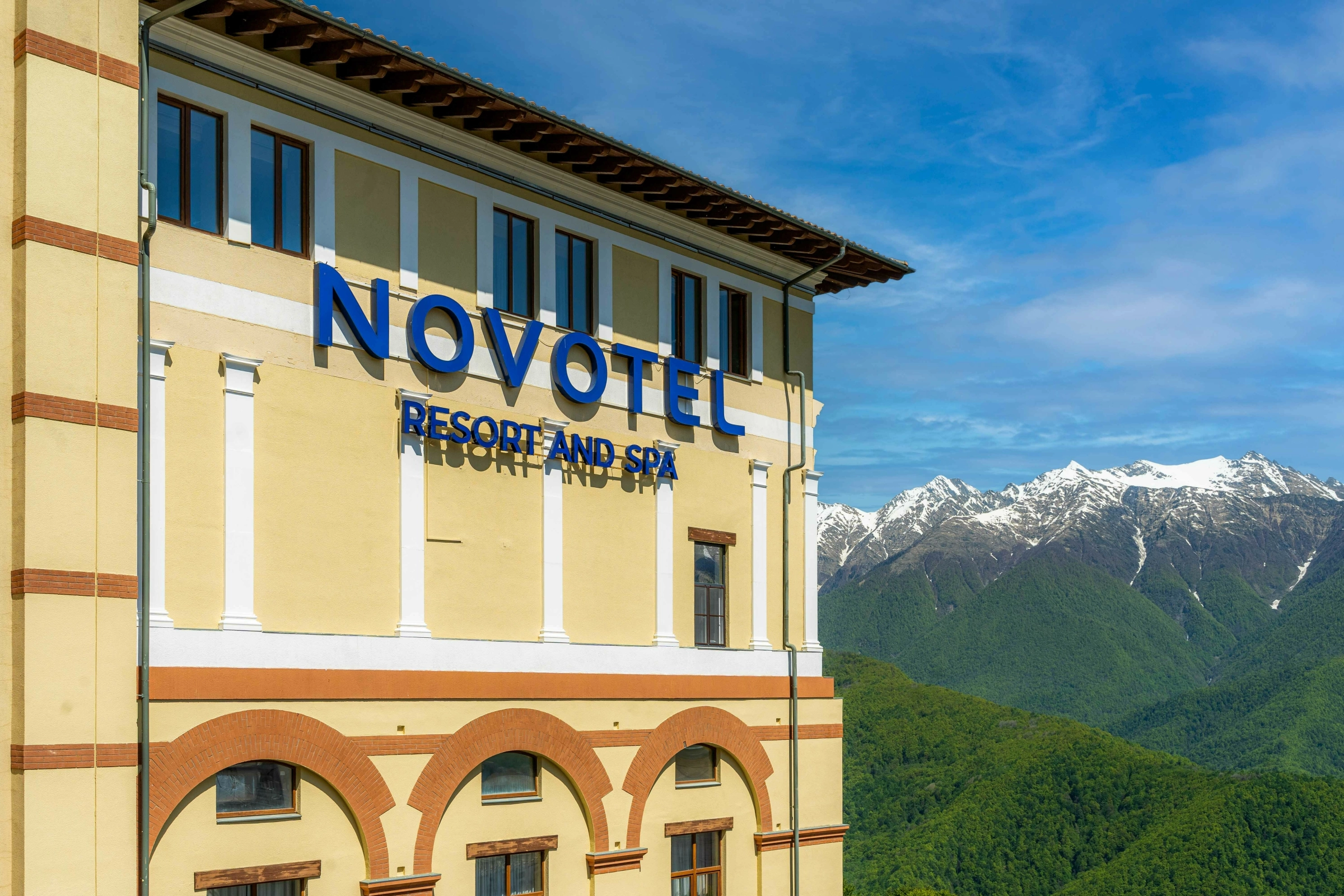 Фасад отеля Novotel с вывеской на фоне зелёных гор и вершин