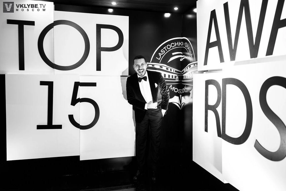 мужчина в смокинге весело смеётся у стенда премии Top 15 Awards