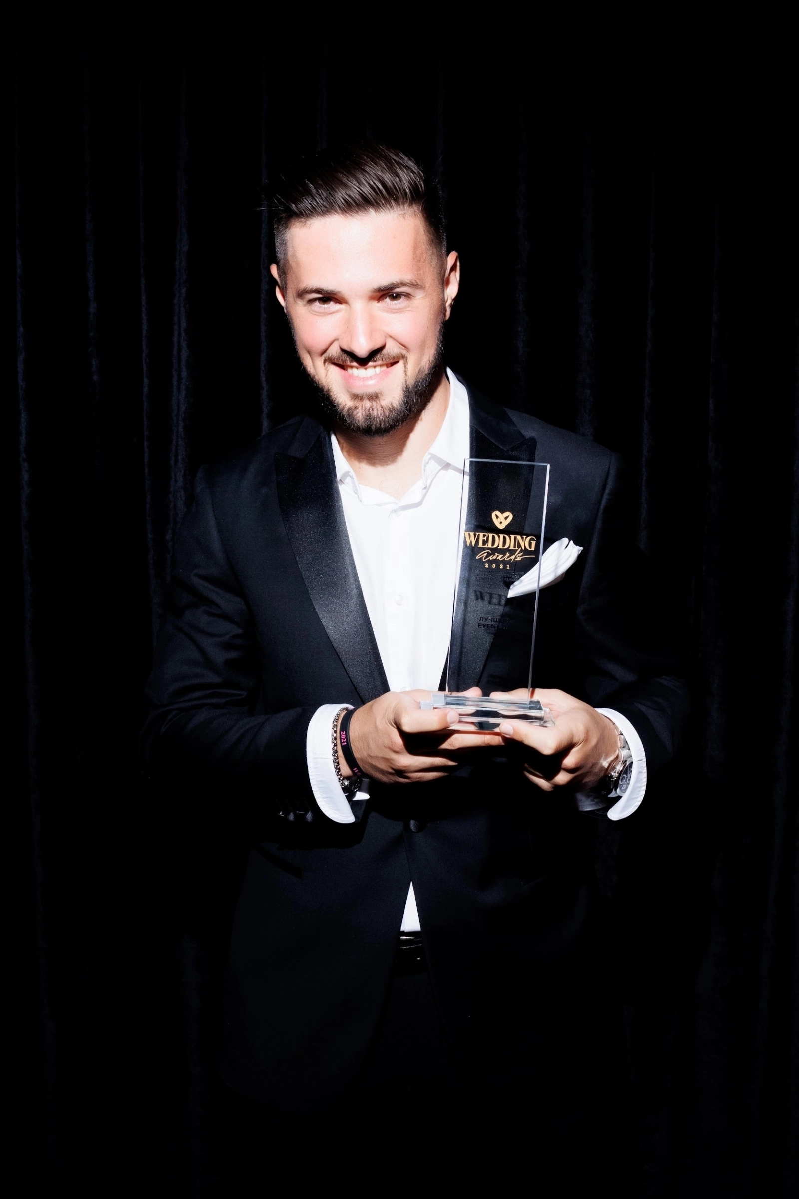 Диджей в смокинге держит награду Wedding Awards на тёмном фоне