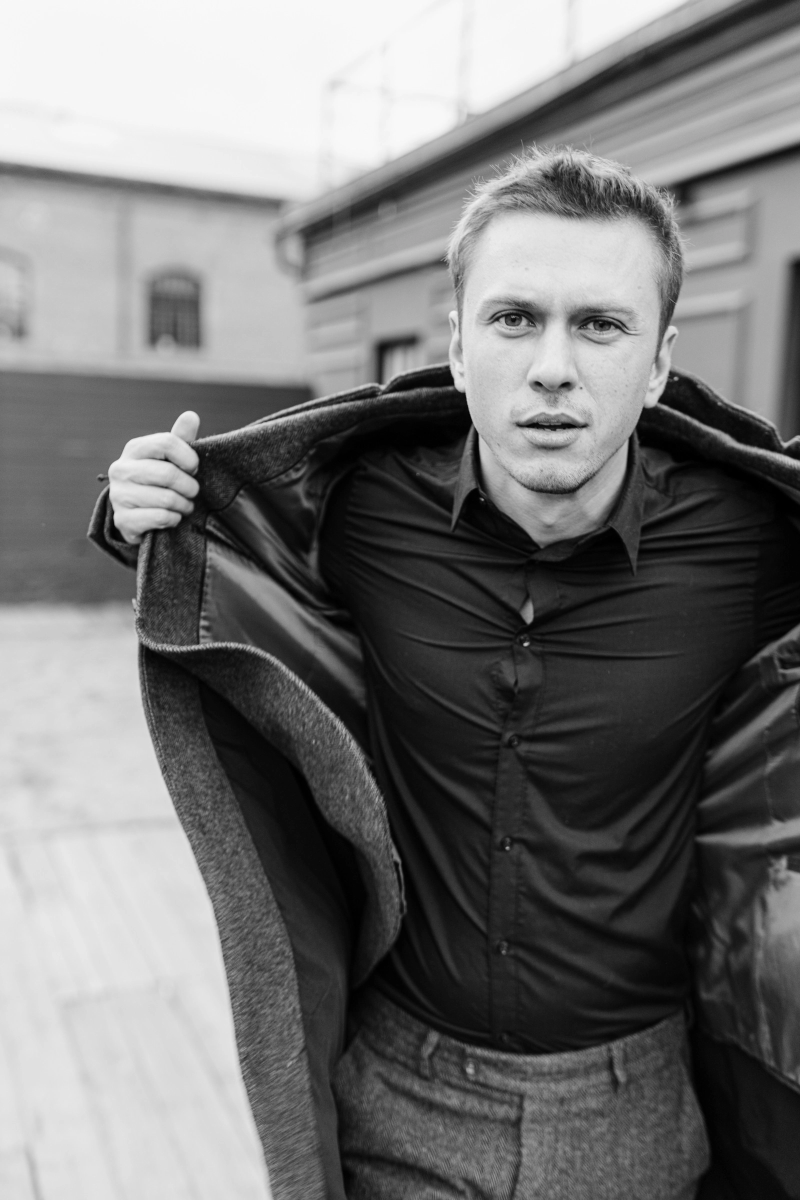 Мужчина в чёрной рубашке держит полы длинного пальто на плечах