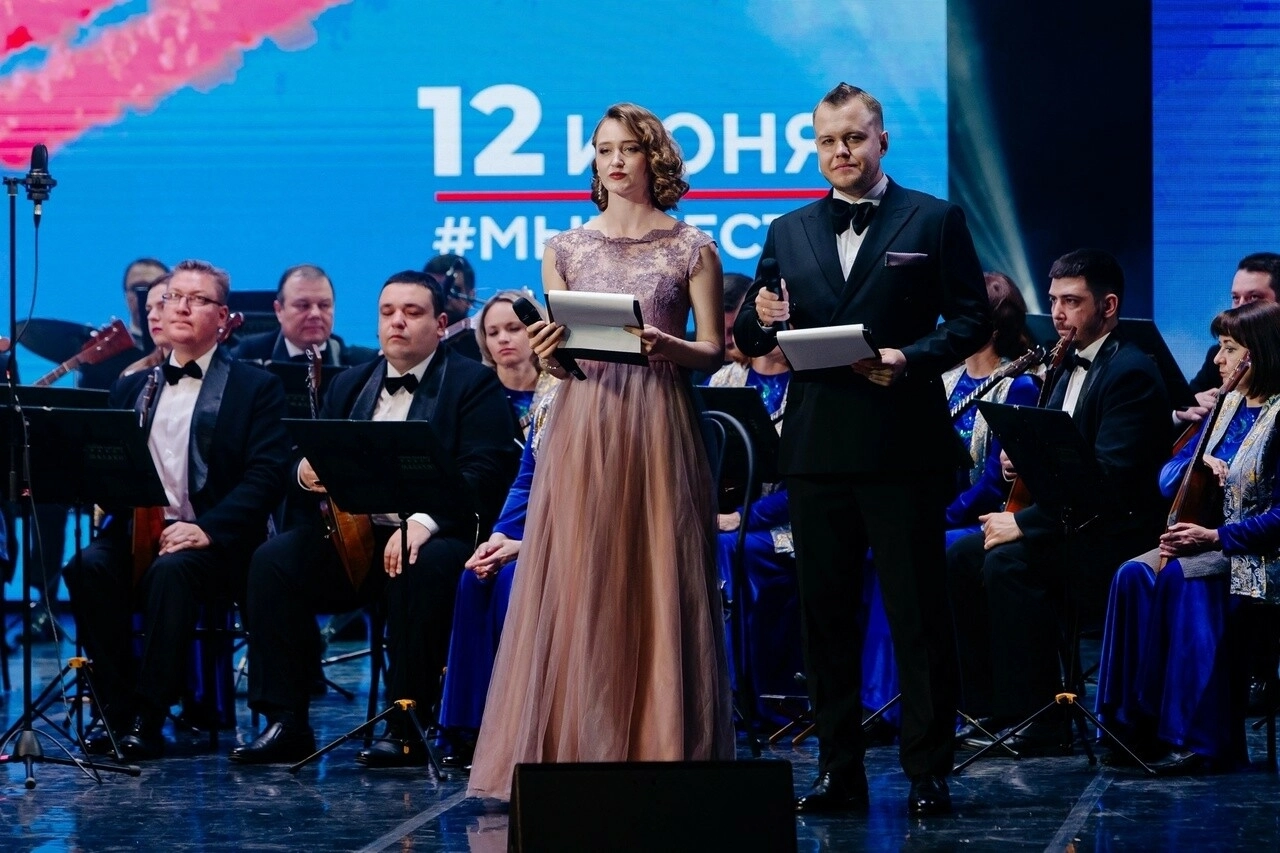 Мужчина и женщина с листами читают текст перед оркестром на сцене