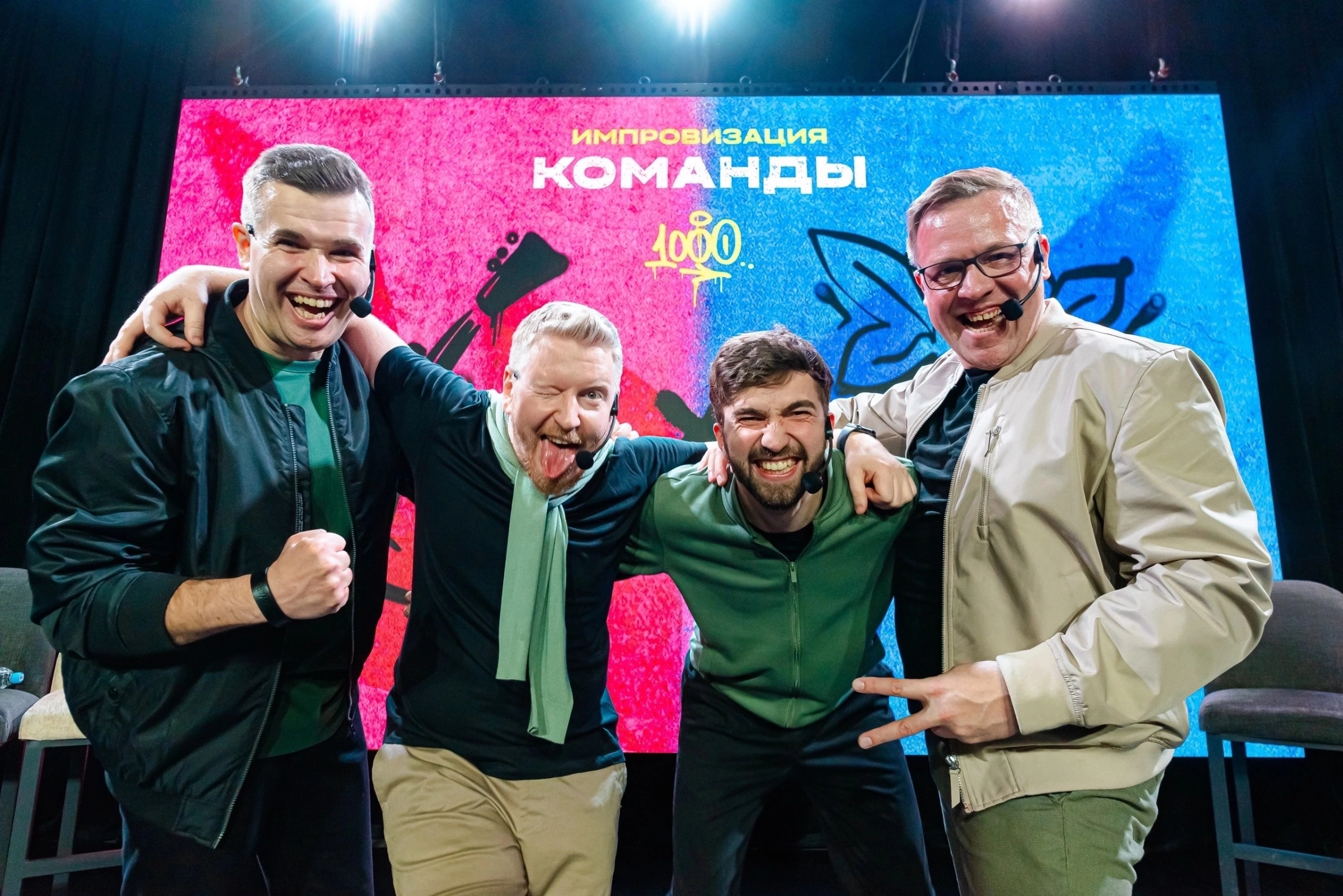 четверо ведущих в куртках обнимаются на сцене шоу-программы с экраном