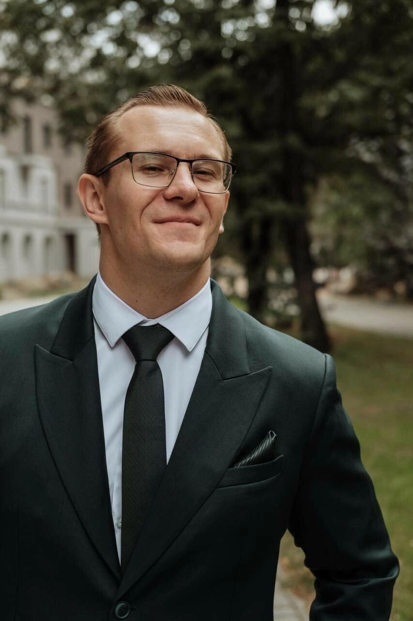 Мужчина в очках и пиджаке улыбается на фоне зданий города
