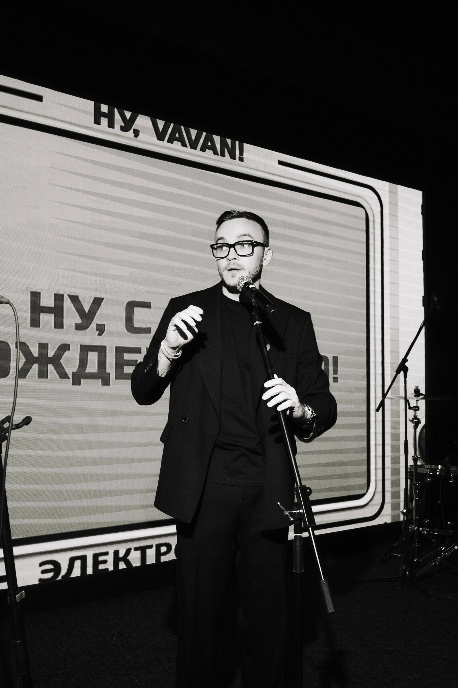 Ведущий в очках с микрофоном на фоне праздничной надписи