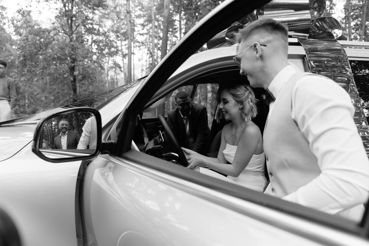 жених и невеста улыбаются в свадебном автомобиле в лесу