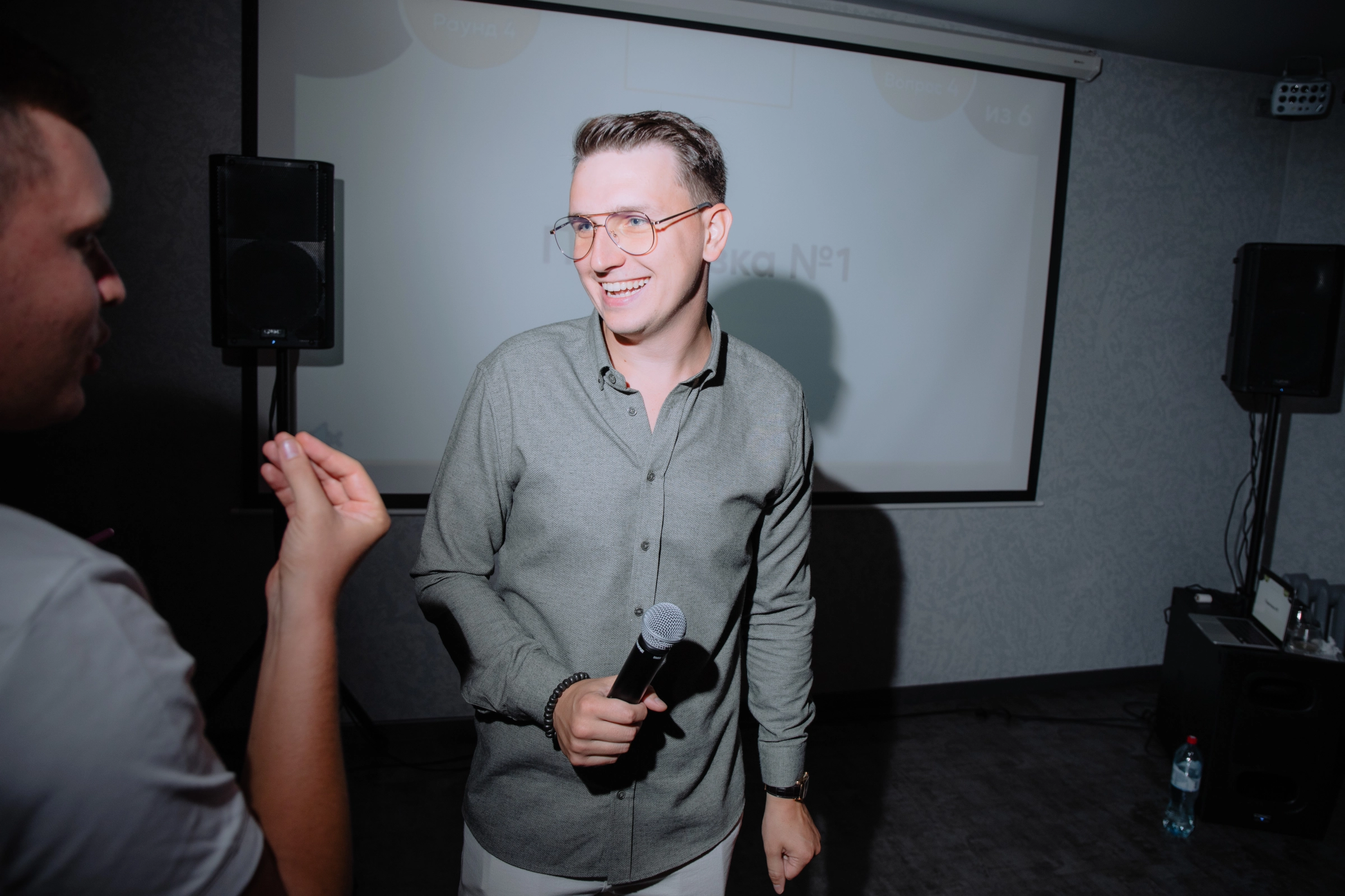 Ведущий улыбается с микрофоном на фоне проекционного экрана