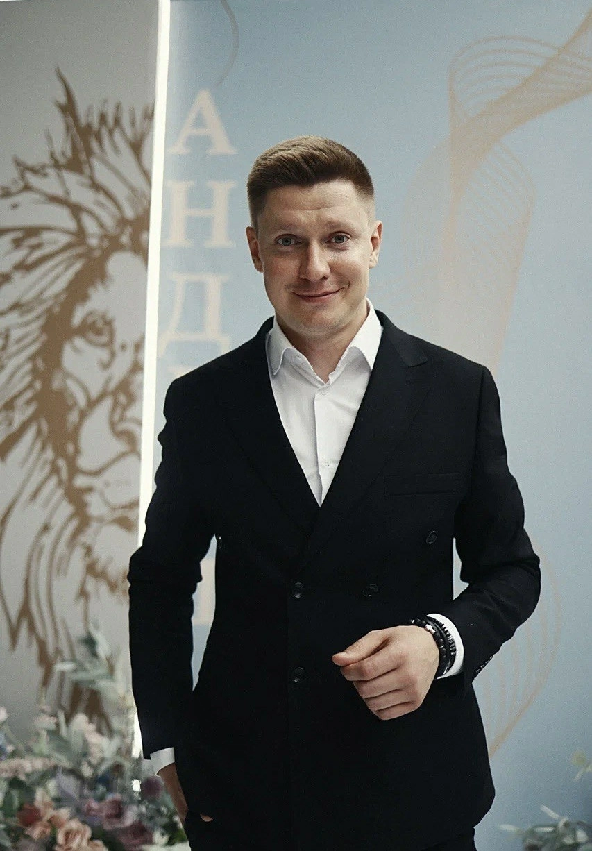 ведущий в двубортном пиджаке позирует на фоне баннера с надписью