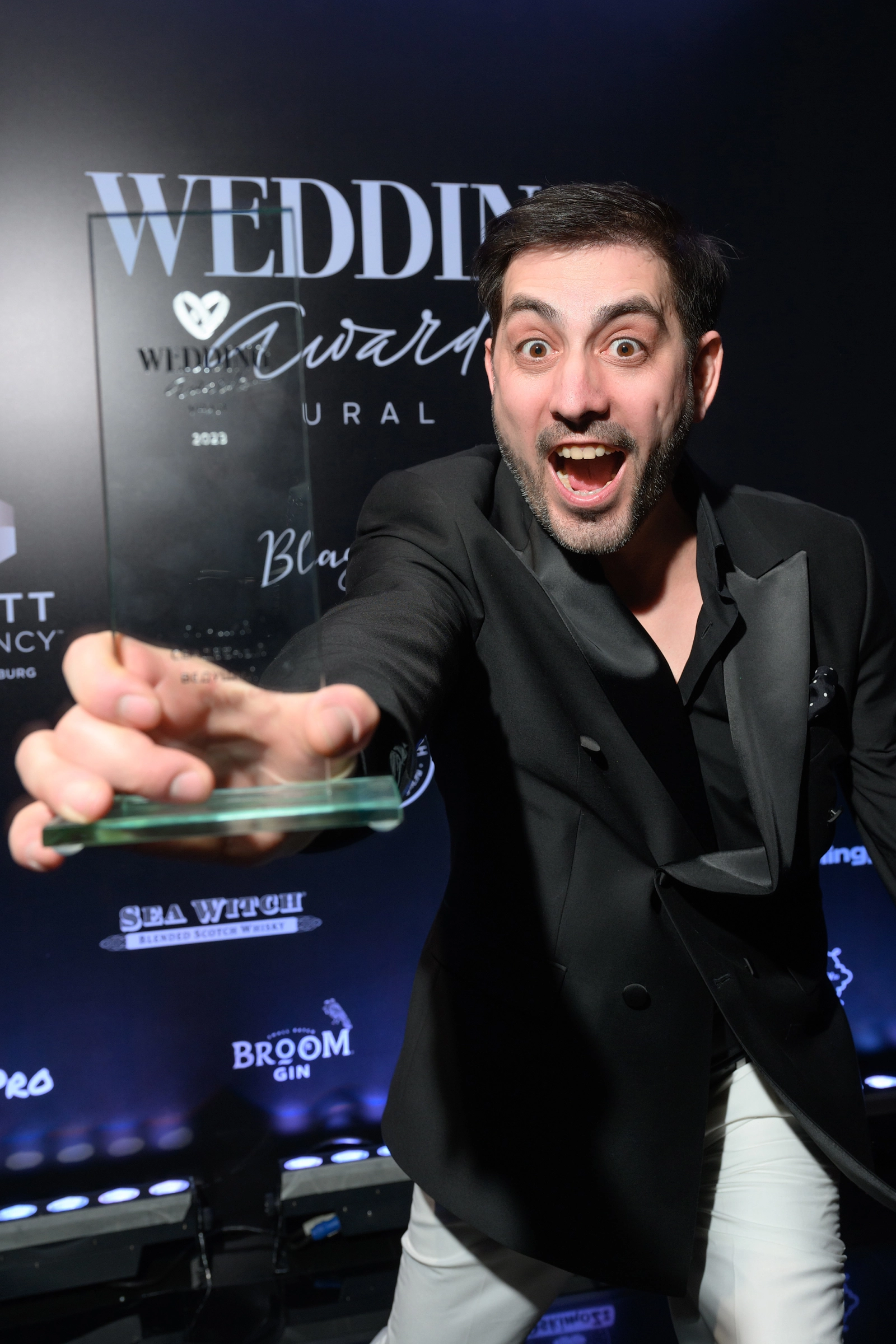 ведущий держит стеклянный трофей премии Wedding Awards на фоне стенда