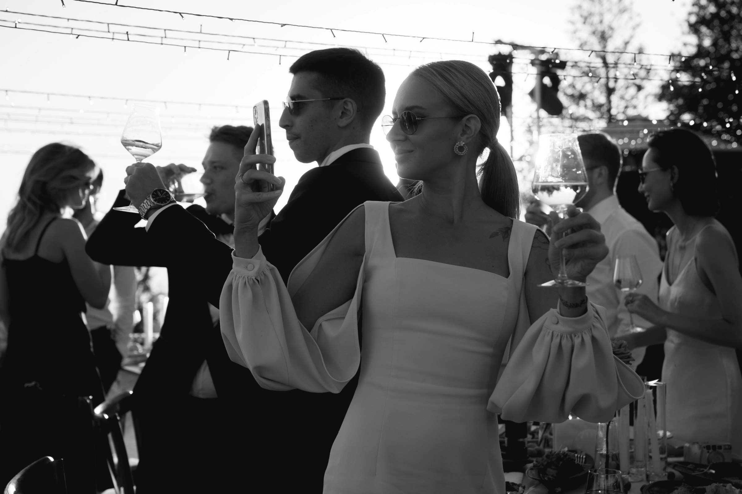 гостья в белом платье с бокалом вина фотографирует вечер на телефон