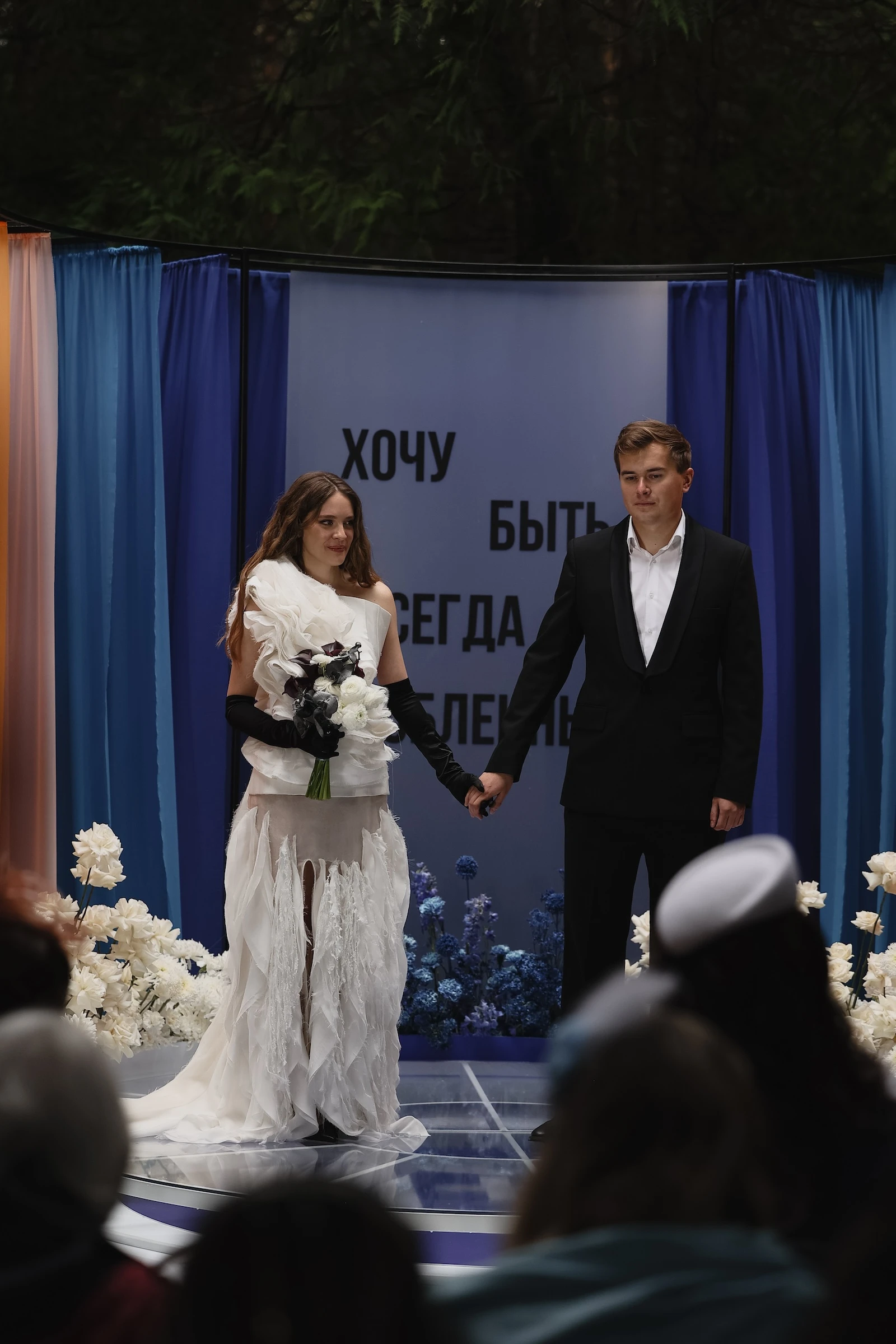 невеста в асимметричном платье и жених держатся за руки на подиуме перед гостями