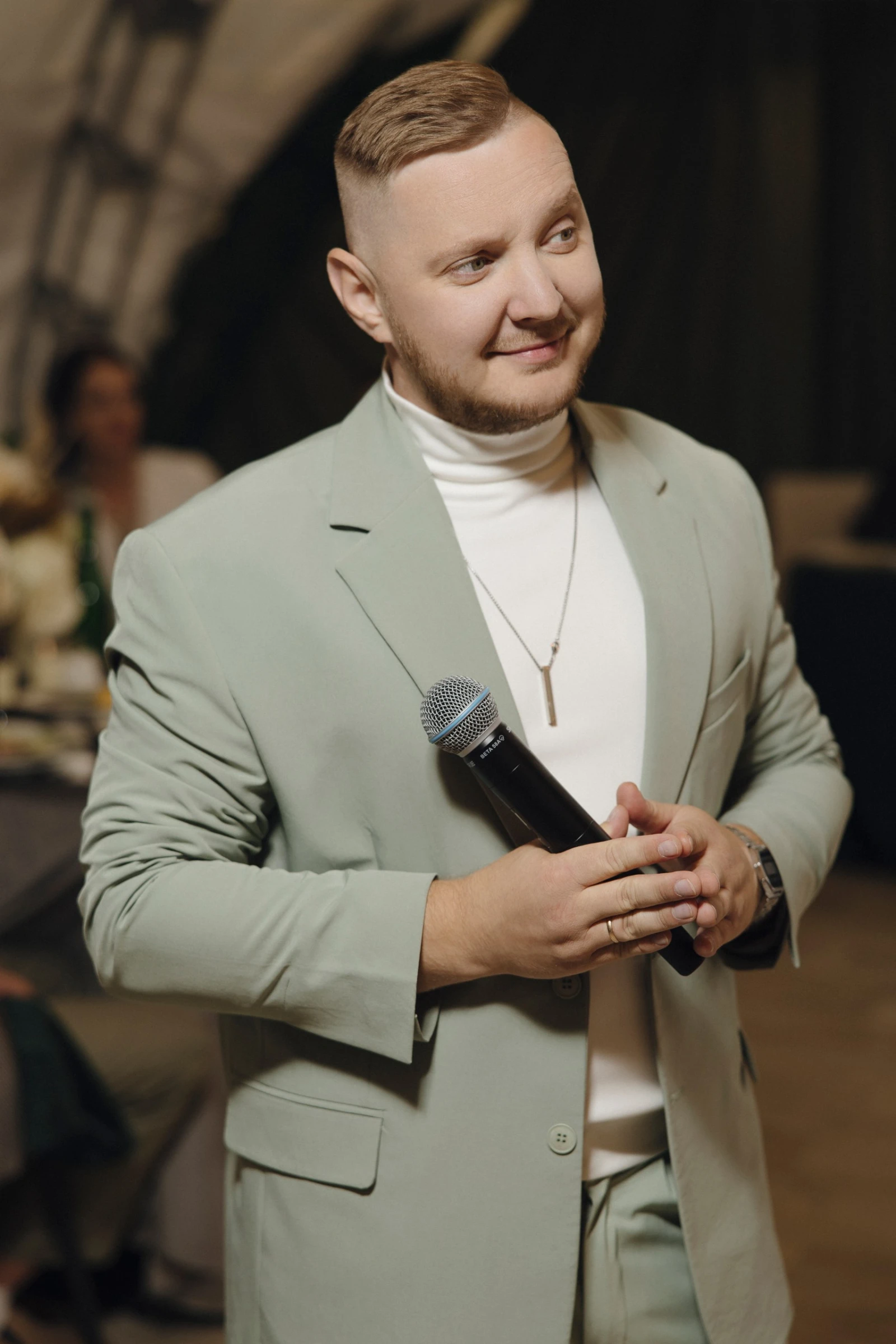 ведущий в мятном пиджаке с микрофоном улыбается на фоне тёмного шатра