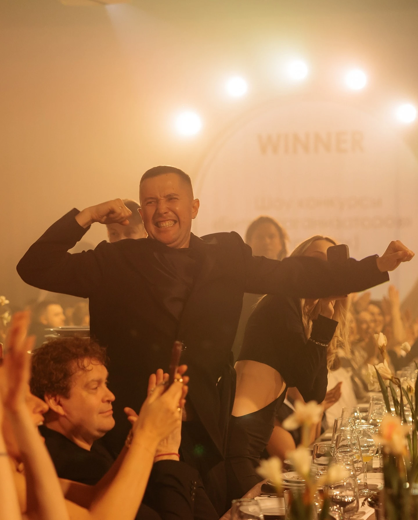 Ведущий радостно поднимает руки у стола на фоне надписи WINNER