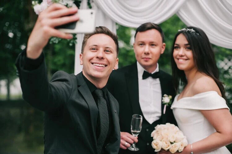 ведущий свадьбы фотографируется с молодожёнами на фоне арки с драпировкой