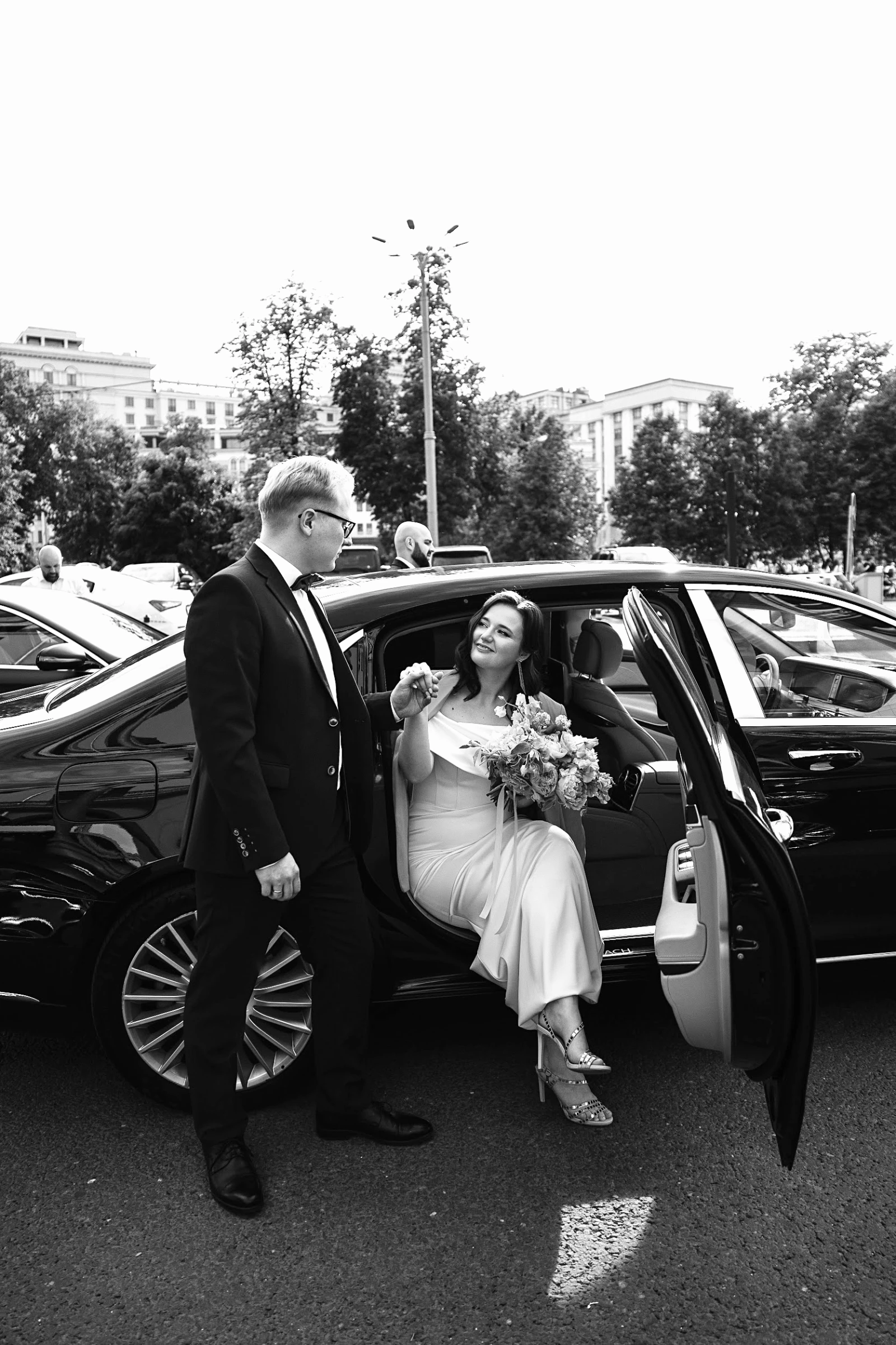 невеста с букетом выходит из автомобиля, жених подаёт руку на улице