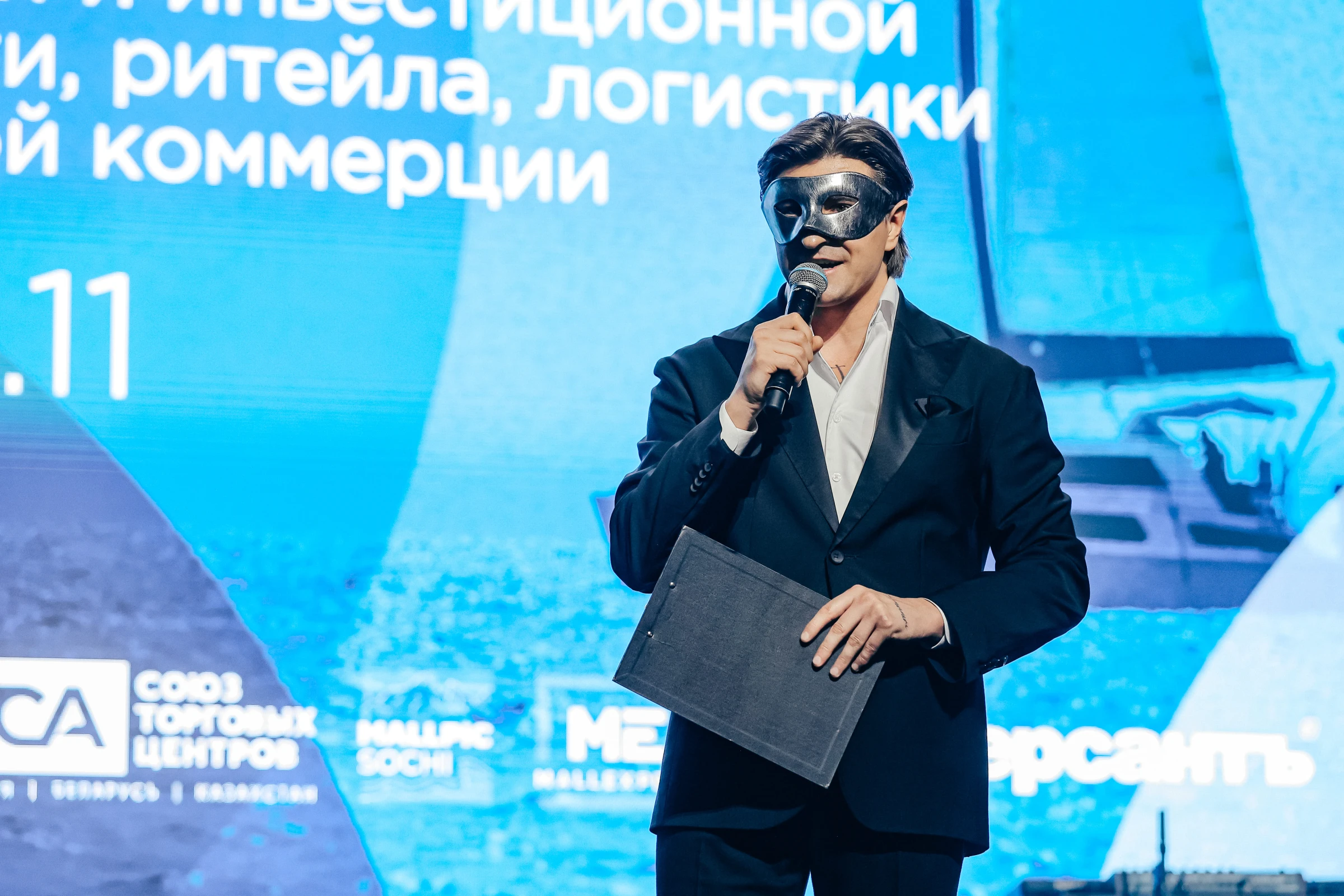 спикер в маске держит папку и микрофон на фоне синего экрана