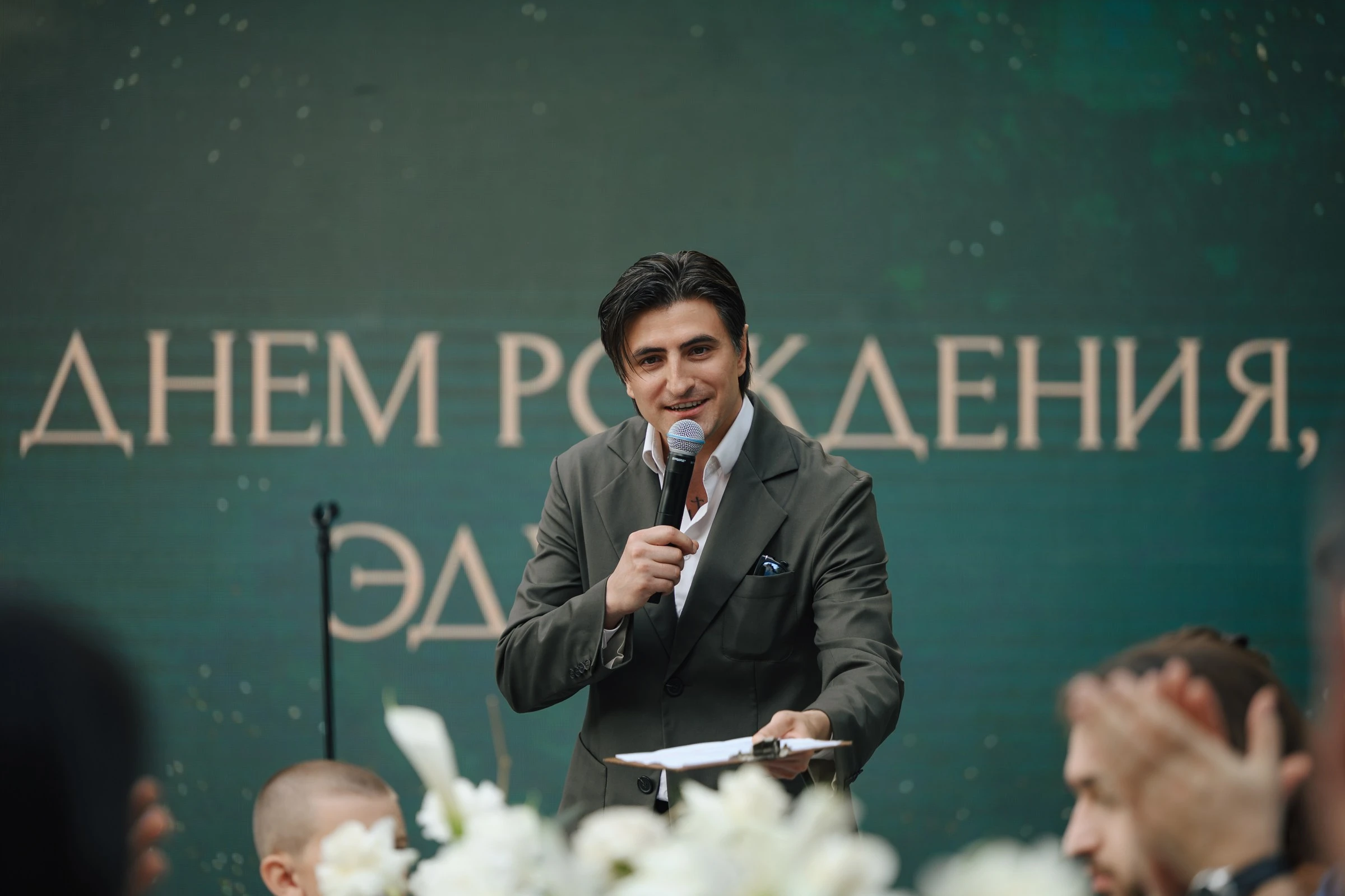 мужчина с микрофоном произносит тост на фоне праздничного баннера