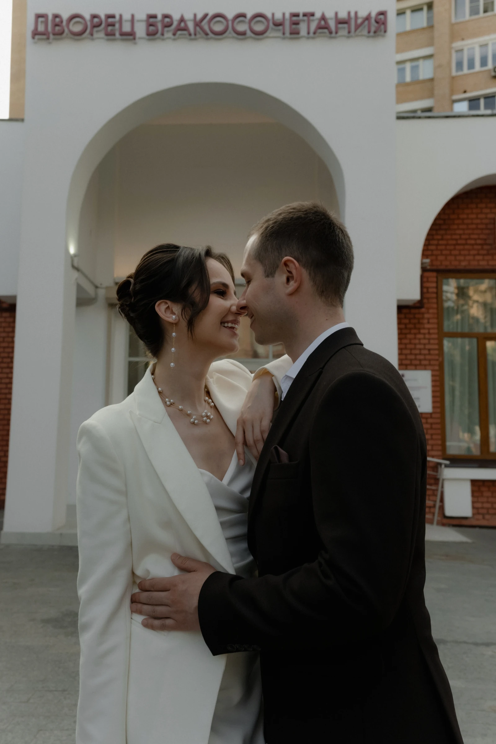 жених и невеста нос к носу улыбаются у здания загса