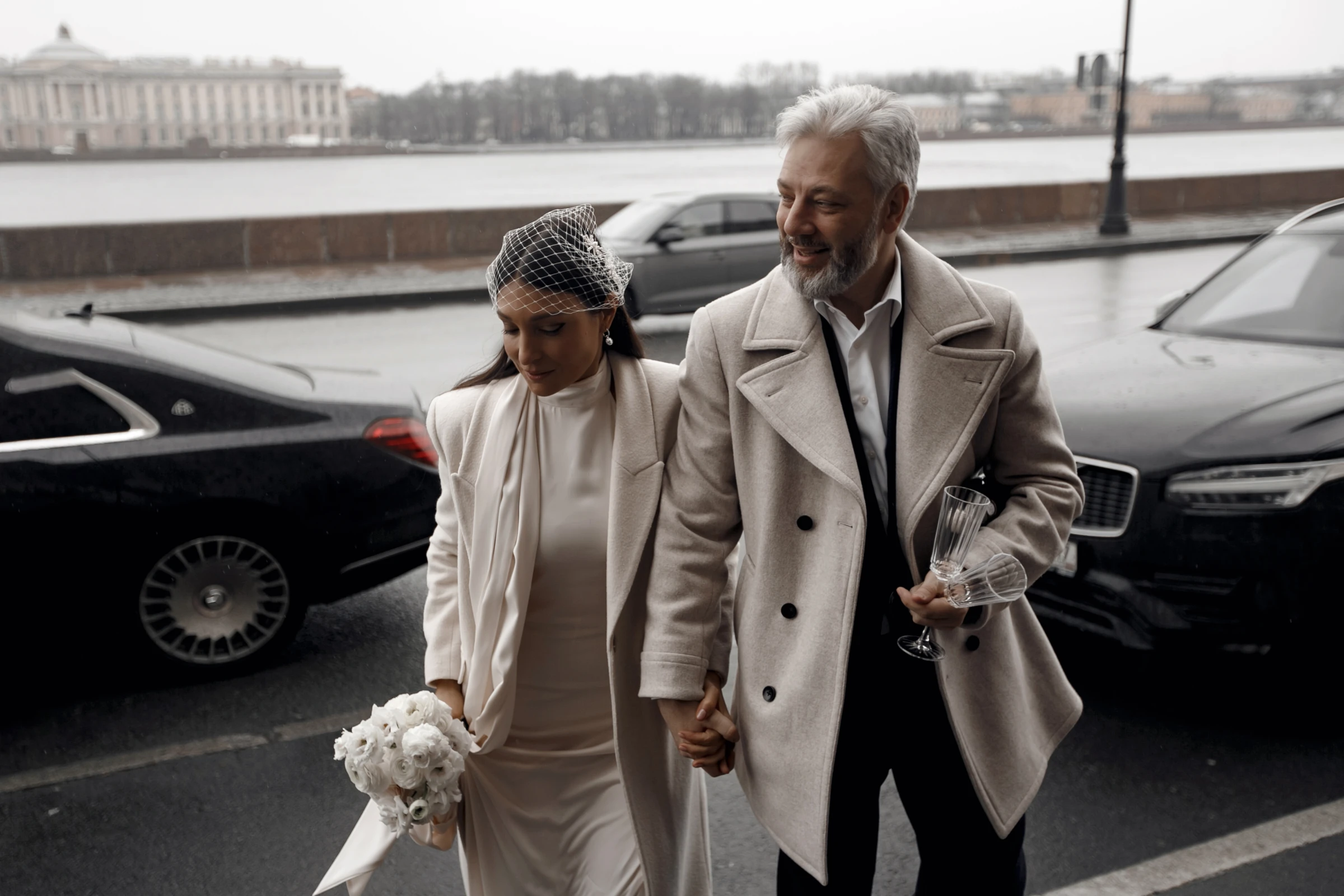 невеста в вуалетке и жених в пальто идут вдоль набережной реки