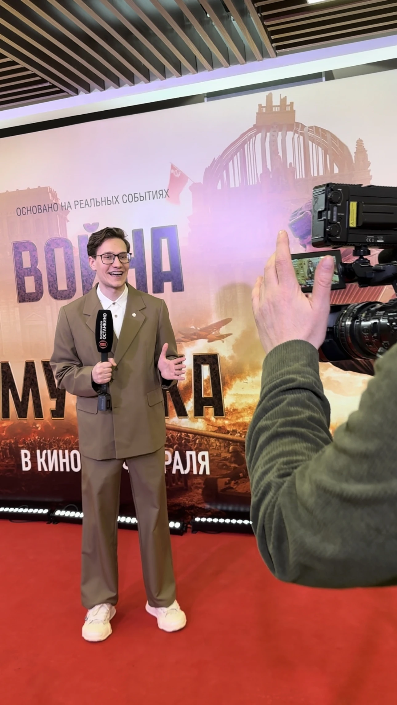 Ведущий с микрофоном стоит на красной дорожке перед камерой