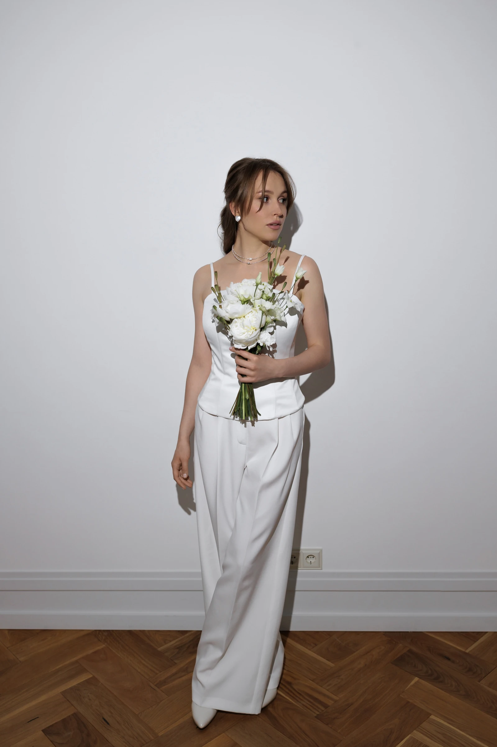 Невеста в белом брючном костюме держит букет белых цветов у стены