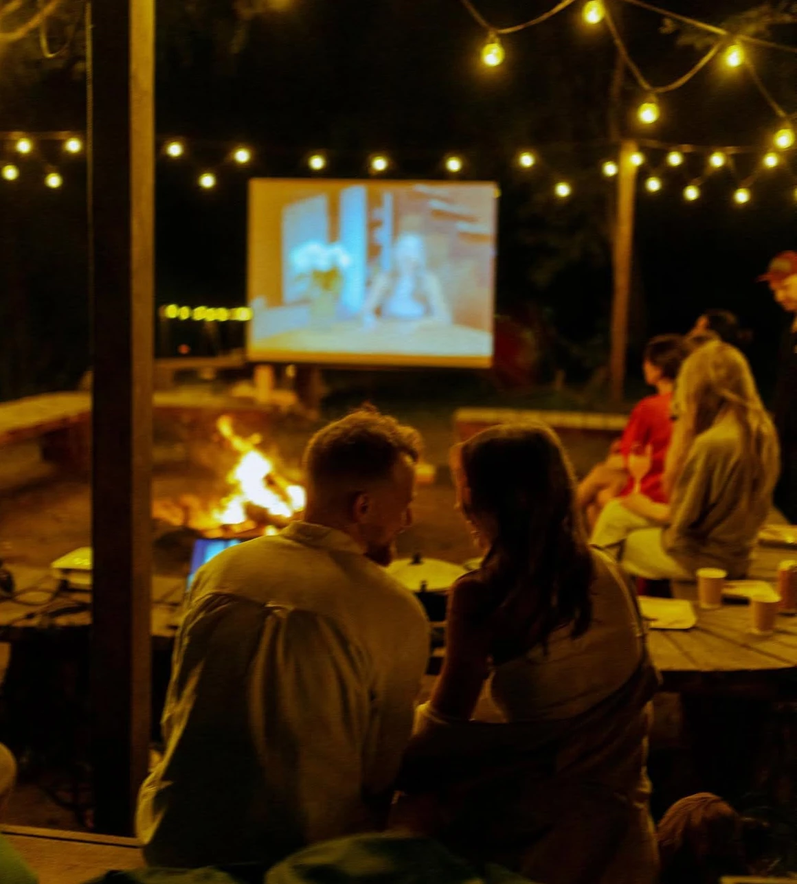 гости у костра смотрят слайд-шоу на большом экране вечером