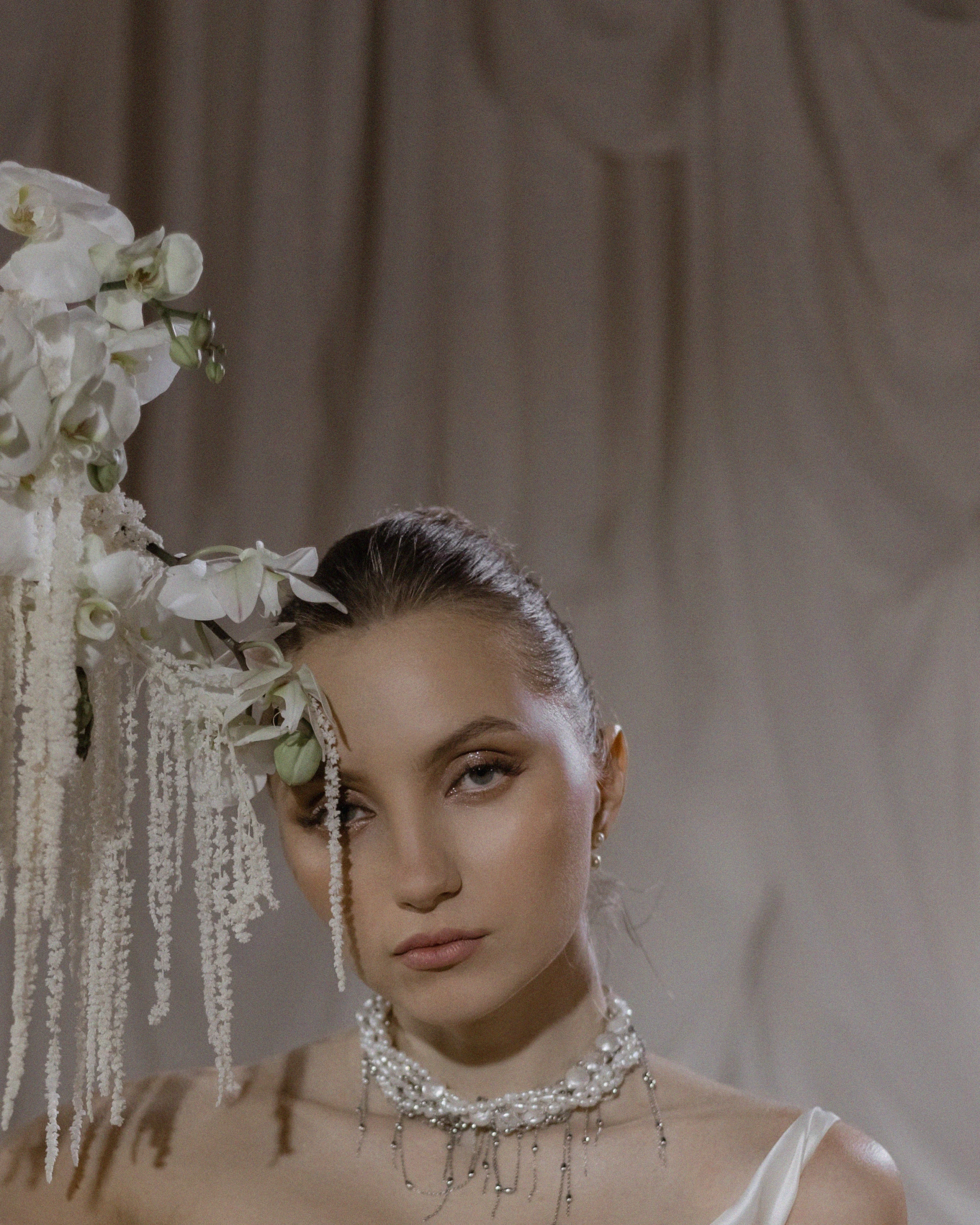 Невеста с жемчужным колье позирует рядом с белыми орхидеями на бежевом фоне