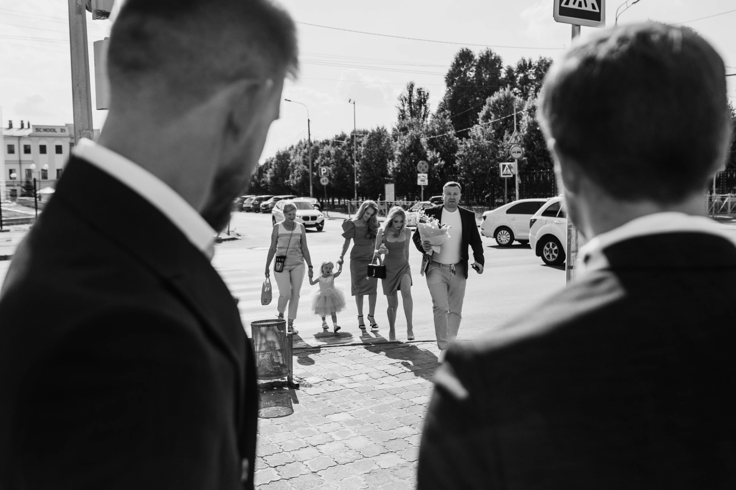 Мужчины в костюмах смотрят на семью с цветами, идущую по городской улице
