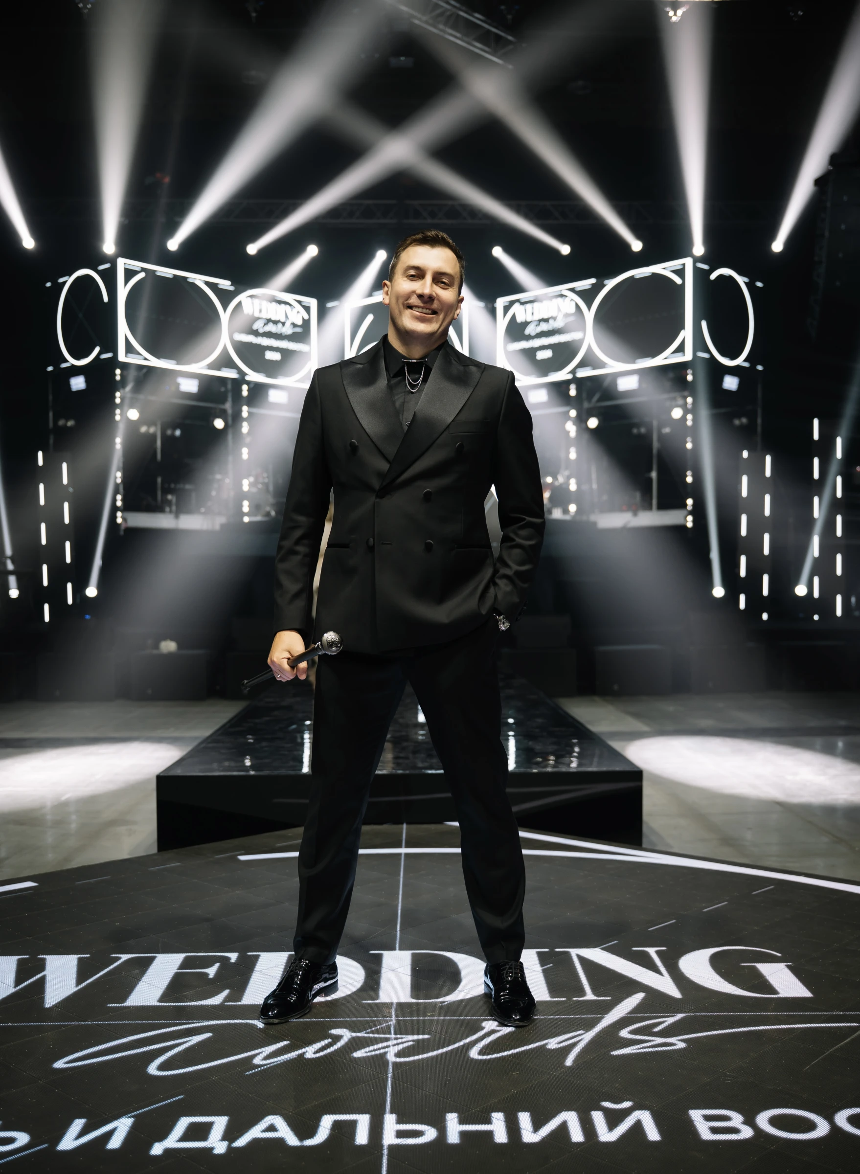 Ведущий в чёрном костюме с микрофоном на сцене премии Wedding Awards